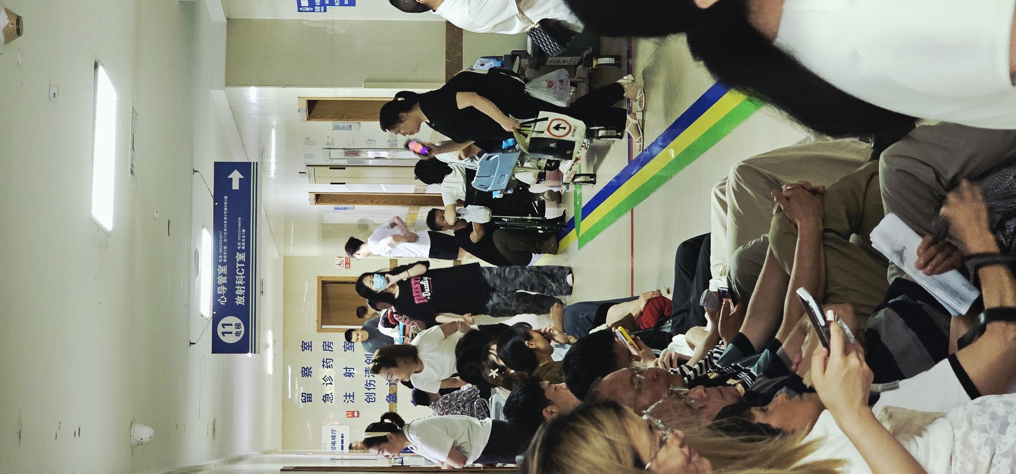 The image depicts a crowded hallway in a medical facility. Many people are sitting and standing, some appearing to be patients or visitors, waiting. Some are using mobile phones. A woman in black is attending to someone in a wheelchair. Signage in Chinese indicates "Cardiology Department" and "Radiology CT room." The sign also includes the phone numbers 18523522017 and 18323149696. Additional signs mention "Observation room," "Emergency Pharmacy," "Injection room," and "Trauma surgery." The location is identified as the city of Yuzhong District, China.