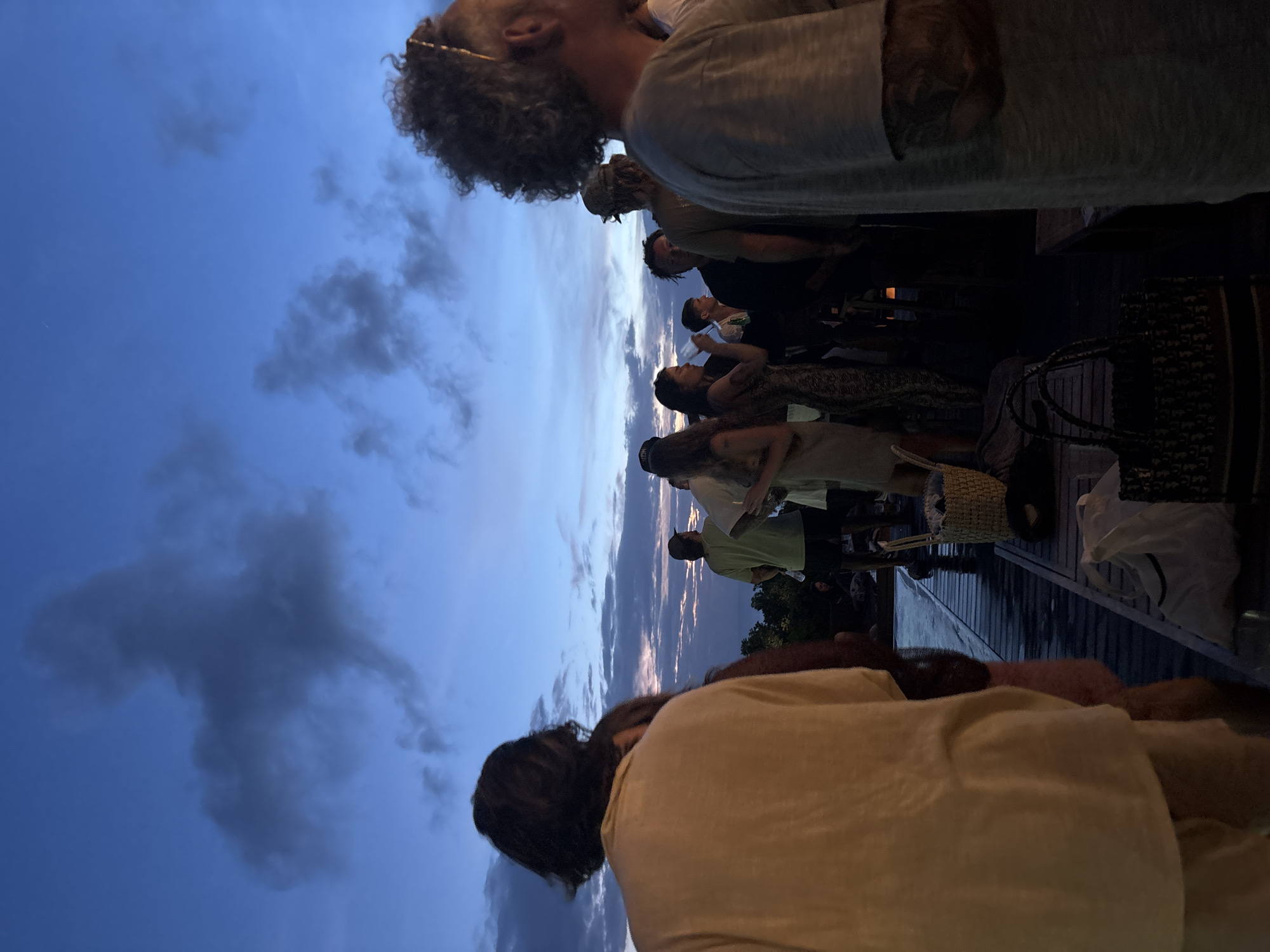The image captures an outdoor gathering of approximately ten to fifteen people on a wooden deck at dusk in Kuta Selatan, Indonesia. The sky dominates the upper portion of the frame, displaying a deep blue overhead that gradually transitions to lighter blues, purples, and faint orange hues towards the horizon, indicative of twilight. Scattered grey and wispy clouds are visible across the sky.
The individuals are mostly standing, facing generally towards the right (if the image were upright), suggesting they are socializing or observing an event. In the immediate foreground, the back of a person with dark hair and a light-colored top is visible. Further back, a man with curly hair and a light grey shirt is prominent among the group. Other attendees are dressed casually, with some wearing patterned clothing and hats.
The scene includes practical objects such as a woven basket and various bags on the deck. Dimly lit dark structures, possibly parts of buildings or fences, can be seen in the background. The overall mood appears relaxed and social. No visible text is identifiable in the image.