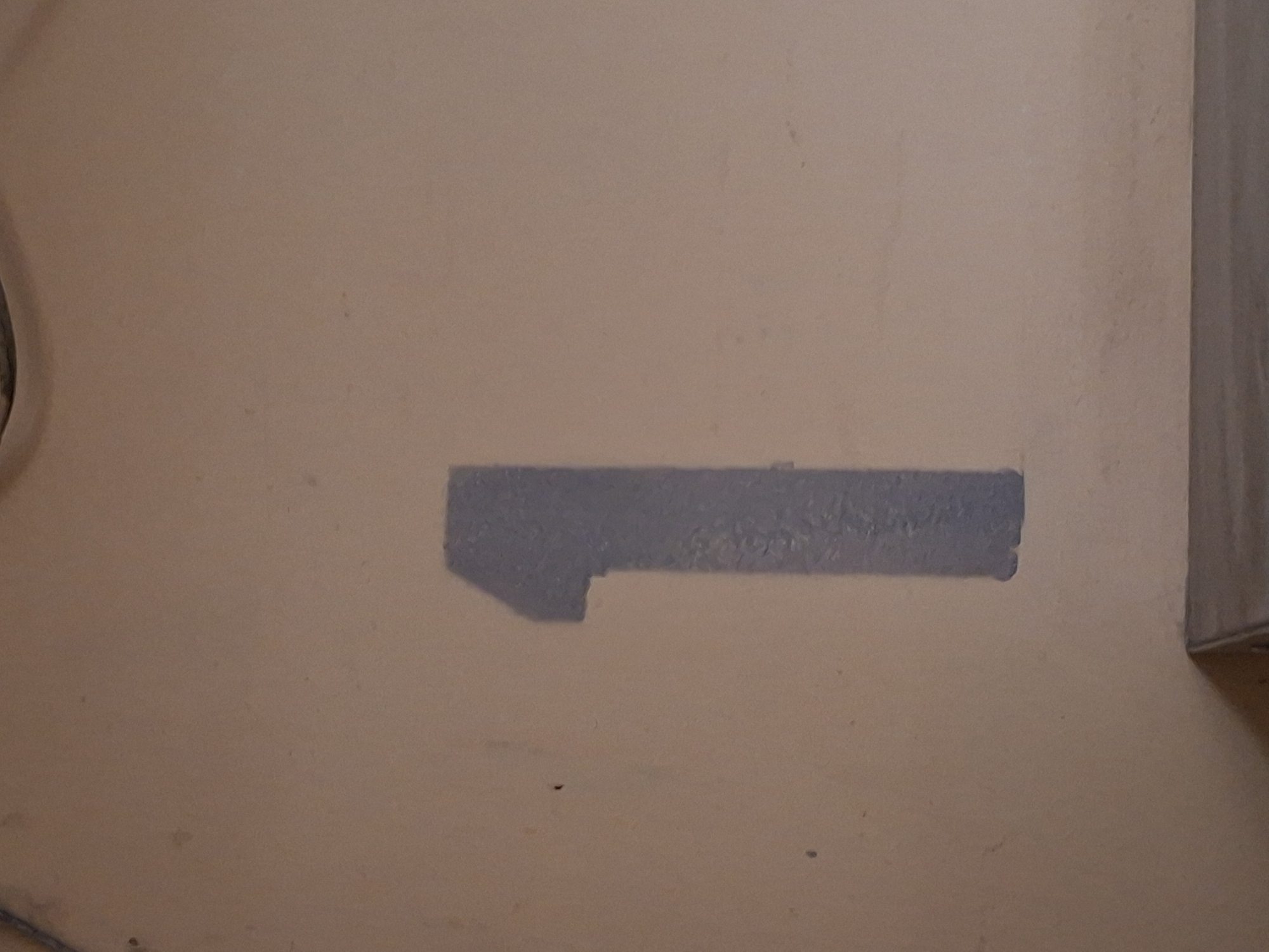 The media file displays a section of a wall, likely indoors, with a light brown or beige painted surface. A noticeable rectangular shape with a notched corner is painted in a muted blue-gray color. The paint appears somewhat textured and possibly applied unevenly, suggesting it might be a patch or a stencil. The wall surface itself has a matte finish and shows some subtle imperfections like small specks and faint marks.
To the left of the wall, a curved, dark object is partially visible, possibly the edge of a piece of furniture or equipment. On the right side of the image, a vertical element with a lighter, wood-toned surface meets the wall at an angle, creating a corner. This suggests the wall is part of a room or an interior space.
There is no visible text in the image. The lighting appears to be soft and even, not indicating a specific time of day or strong directional light source. The overall scene is static and focuses on the detail of the painted mark on the wall. The context of Kazan, Russia, provides a geographical location but no specific visual cues within the image confirm this.