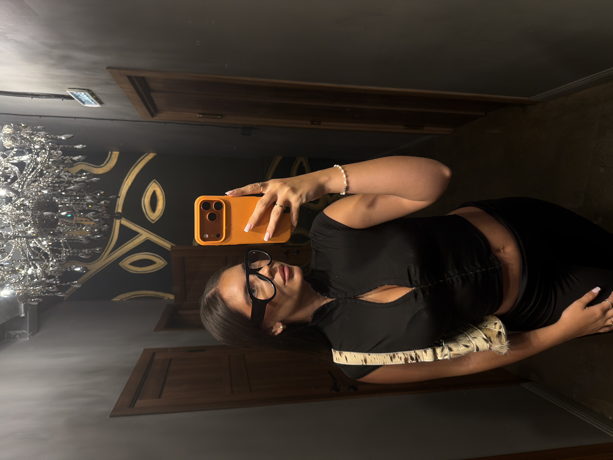 The image shows a woman taking a selfie in what appears to be a stylish interior. She is wearing glasses and a black outfit, and is holding an orange phone. A chandelier is visible in the background, along with ornate wall decorations. The setting seems to be indoors, possibly in a hotel or a high-end establishment. The city of Krasnoyarsk, Russia, cannot be determined from this image alone.