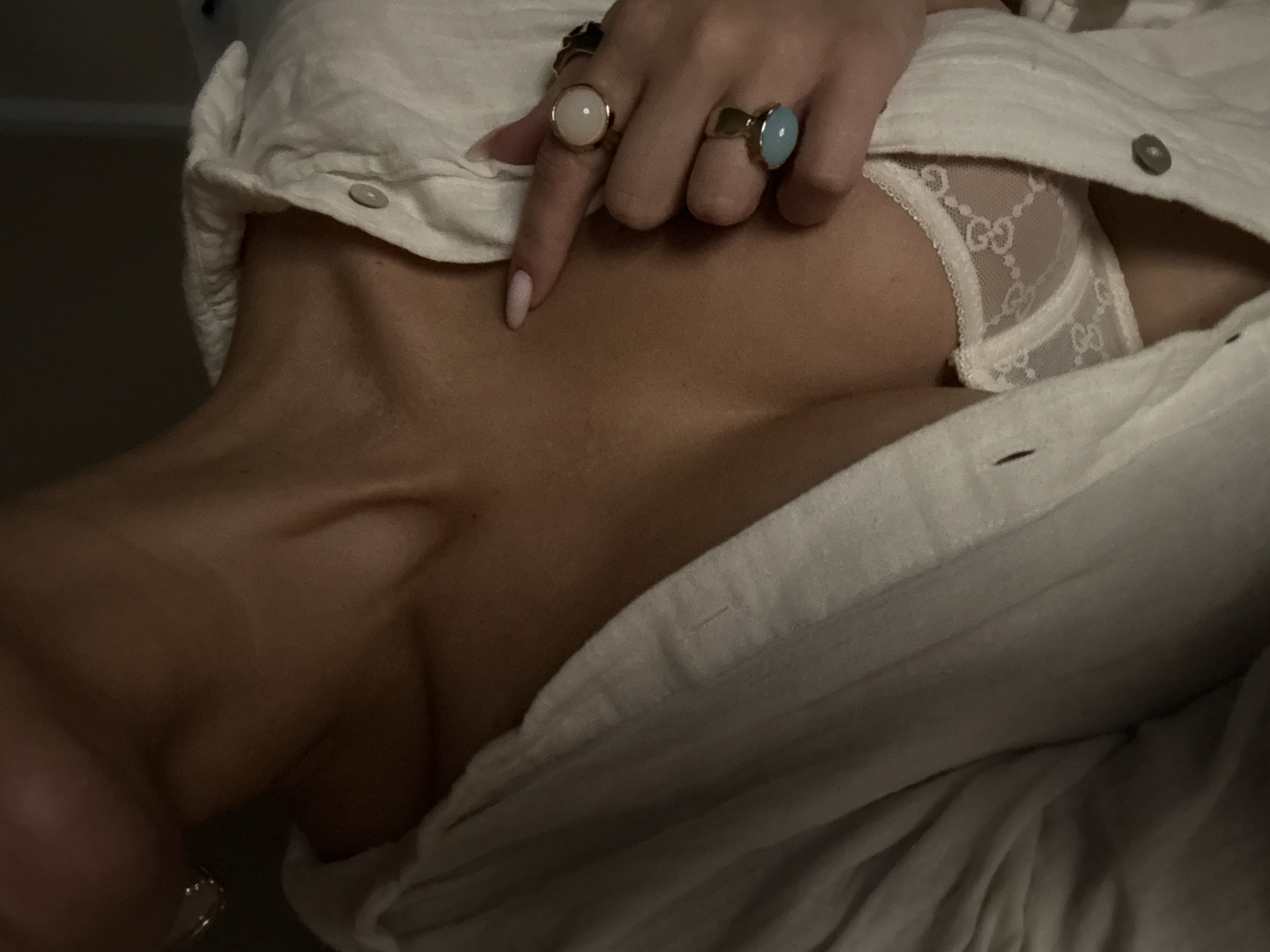 A low-angle close-up image shows a woman's torso, neck, and upper chest. She is wearing a partially unbuttoned light-colored shirt or blouse that exposes a white lace undergarment. The woman's left hand is visible, adorned with multiple rings including one with a large white stone and another with a blue stone. The skin appears to be lightly textured. The location, Chaikovsky, Russia, is not evident in the image. The lighting is low, creating shadows and depth.