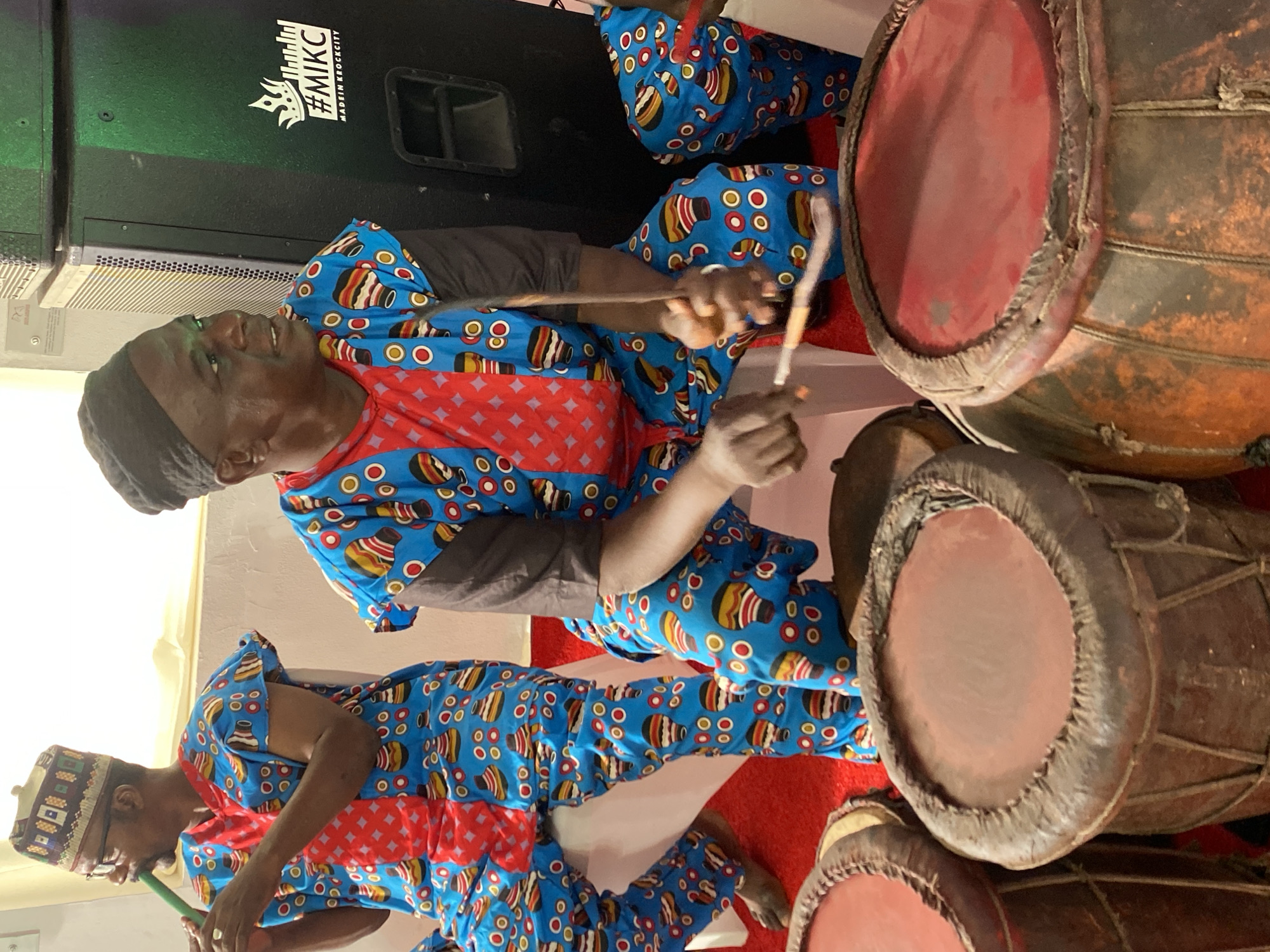 The image shows two men playing drums in Kwarau, Nigeria. The men are wearing matching blue patterned outfits with red geometric designs. One man wears a black head covering, while the other wears a patterned cap. They are seated next to a set of three drums. The drums are brown and have red drumheads. A large black speaker with white writing is visible in the background. A white wall is also visible behind the speaker. The setting appears to be indoors, with a red and white rug.