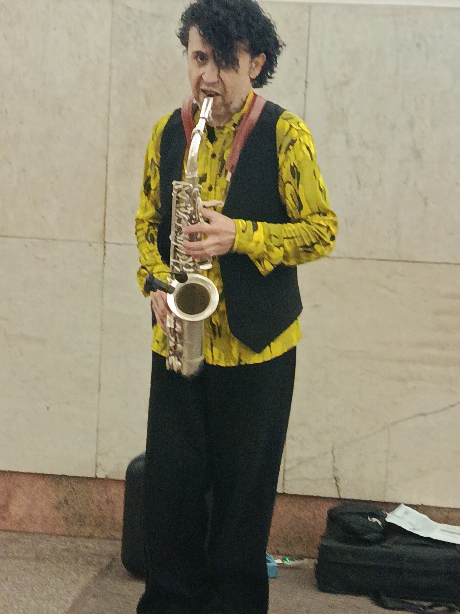 A man is standing and playing a saxophone in an indeterminate indoor location. He wears a yellow, long-sleeved shirt with a black pattern, a black vest, and black pants. He has dark curly hair. He holds a silver saxophone with a red strap, and his mouth is on the mouthpiece. A black bag sits on the floor next to him. The background is a wall with beige panels. The city is Unknown, Unknown.