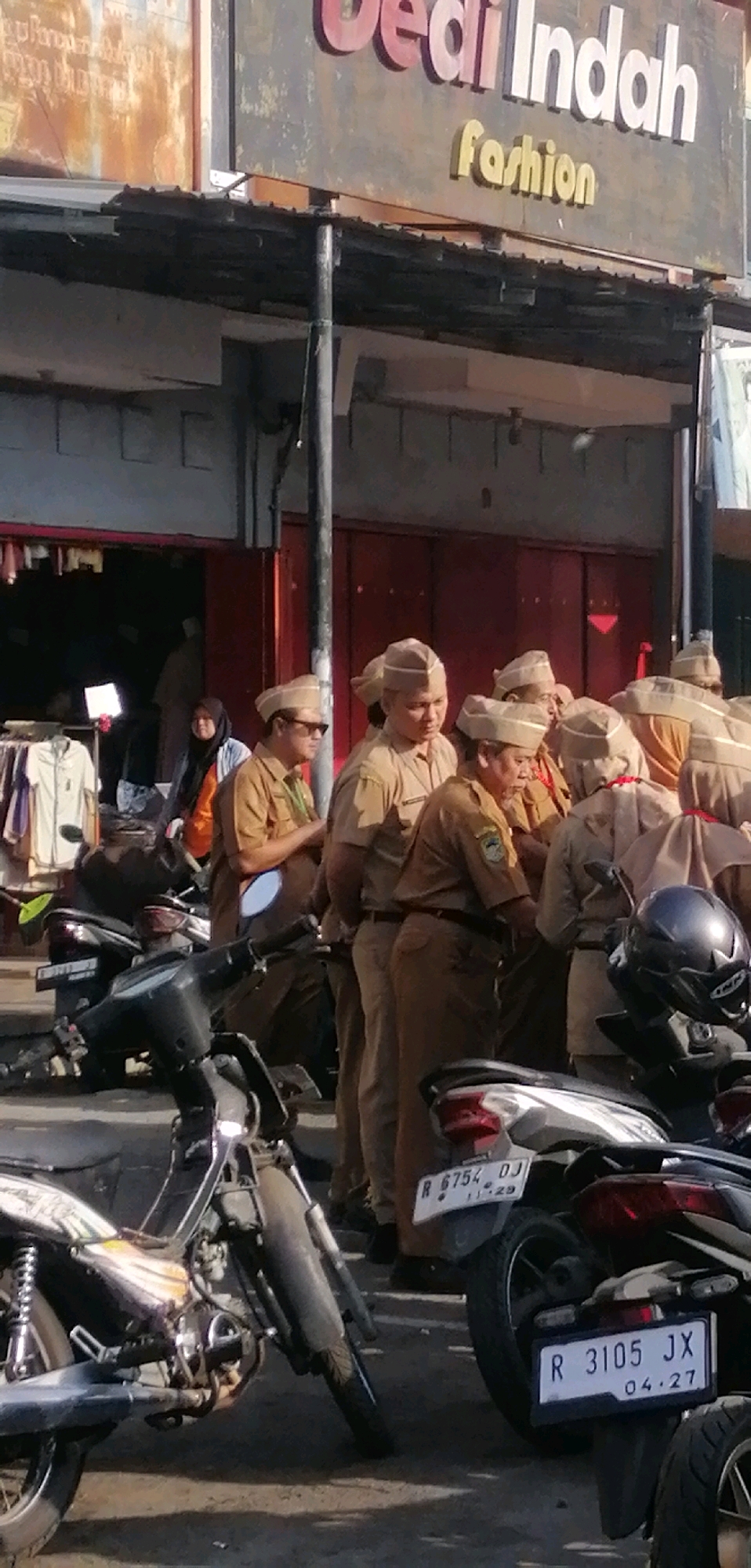 The image depicts an outdoor scene in Ajibarang, Indonesia, likely in front of a retail establishment identified by a sign reading "Gedi Indah fashion." In the foreground, several motorcycles are parked, some with visible license plates. The license plates read "R 6754 DJ" and "R 3105 JX." The environment appears to be a street or market area with a weathered building facade and overhead covering.
A group of people, predominantly men, are the central focus of the image. They are all wearing matching light brown uniforms and caps, suggesting they are part of a uniform group, possibly government employees or attendees of an event. Some individuals are looking in different directions, while others appear to be engaged in conversation or observing something out of frame. One person in the front is wearing sunglasses. Behind the uniformed group, a woman in a hijab is visible, along with racks of clothing, indicating a market or store setting. The lighting suggests it is daytime, with the sun casting shadows.