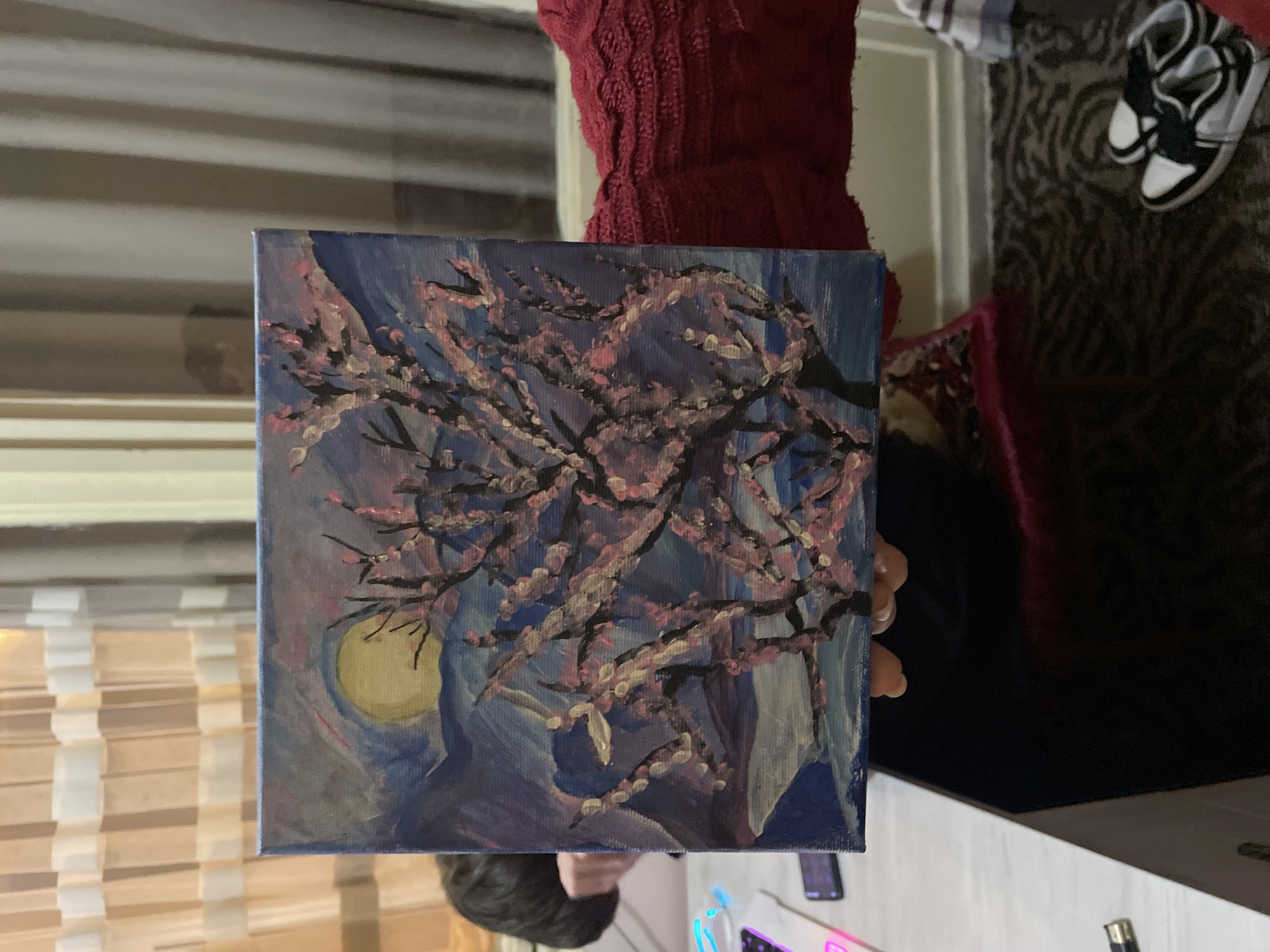 A painting depicting a flowering tree branch silhouetted against a blue background with a yellow moon, is being held. The painting is square and appears to be acrylic on canvas. The background includes horizontal blinds, a person wearing a red sweater, and a pair of black and white sneakers on a patterned surface. The scene is indoors and includes elements such as a white table and a computer keyboard. The location in Yerevan, Armenia, cannot be determined from the image.