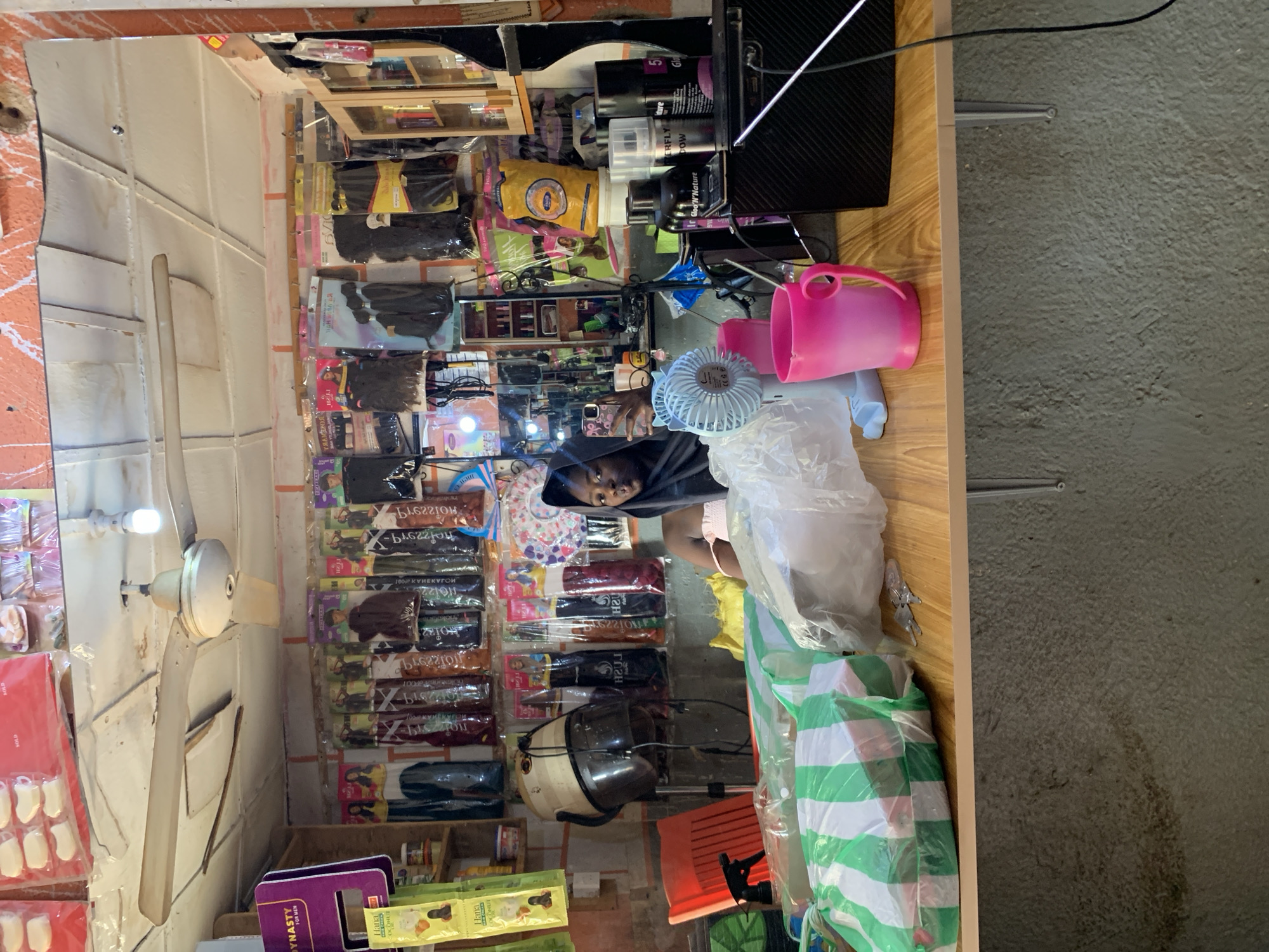 A small shop in Rafin Par, Nigeria, displays numerous packages of synthetic hair extensions on the wall. A young person, possibly a vendor, is present in the shop. Other items visible include a hair dryer, cleaning supplies, a toy fan, a pink cup, and packaged goods. A ceiling fan hangs overhead.