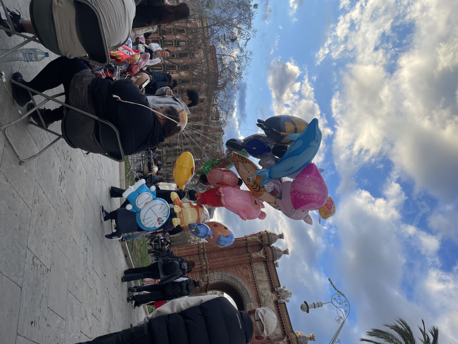 Several people stand near the Arc de Triomf in Barcelona. One person holds many colorful balloons featuring cartoon characters and animals. Other individuals sit in chairs, and a Ferris wheel is visible in the background. The sky is bright blue with scattered clouds, suggesting a sunny day. People wear casual clothing and face different directions. The scene appears to be a busy public area with vendors and tourists.