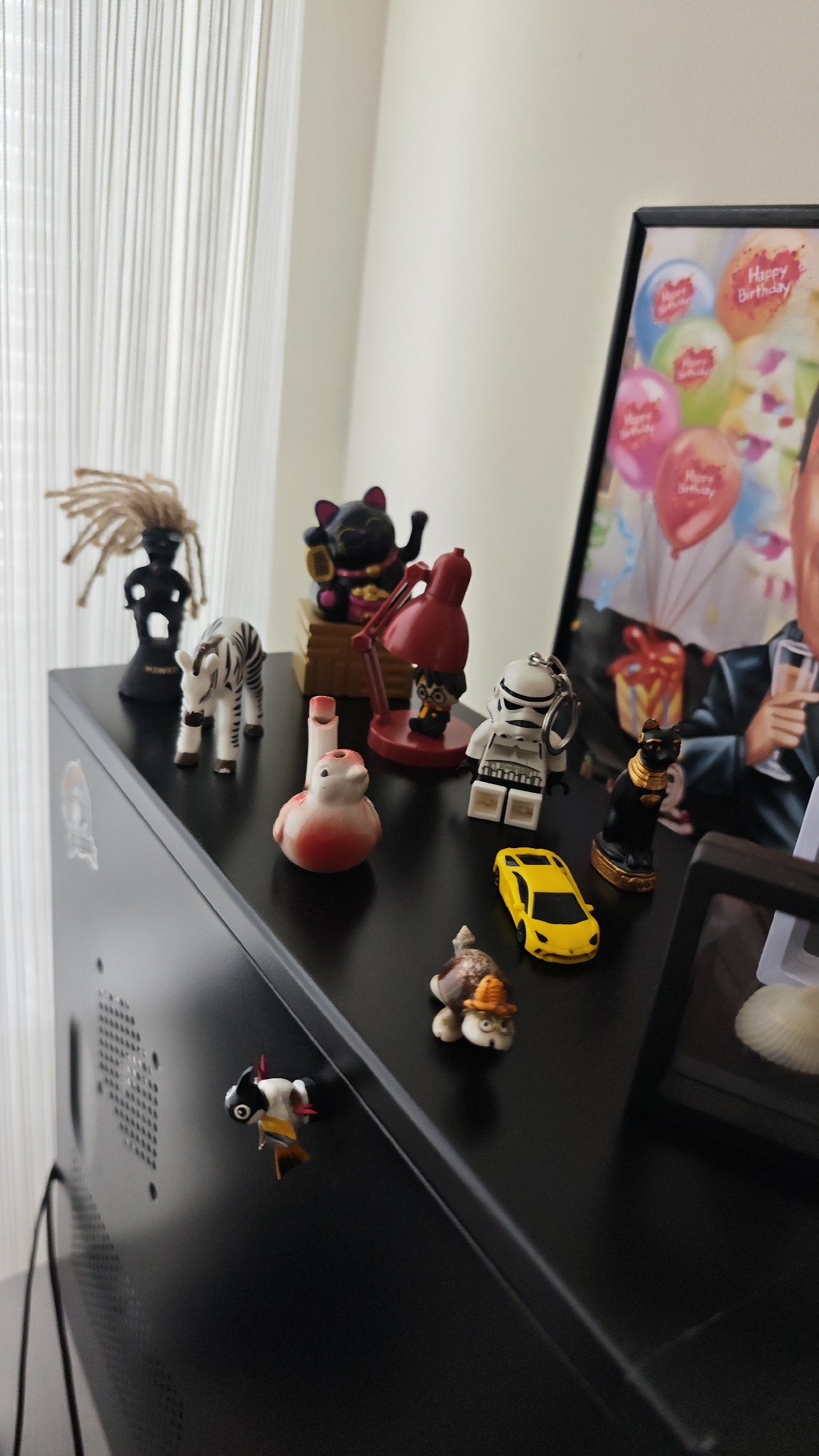 The image shows a collection of figurines and objects arranged on a black surface, likely a TV cabinet or a shelf, in an indoor environment. The background includes white curtains and a picture frame with a photograph showing a person with birthday balloons. The figurines include a black statuette with dreadlocks, a zebra, a maneki-neko, a red lamp, a Harry Potter figure, a Stormtrooper keychain, a red and white bird, a yellow toy car, a turtle with a hat, and a cat statuette. A small airplane figure is also visible attached to the edge of the surface. The environment suggests a domestic setting. It is not possible to determine the location of the image with the information provided.