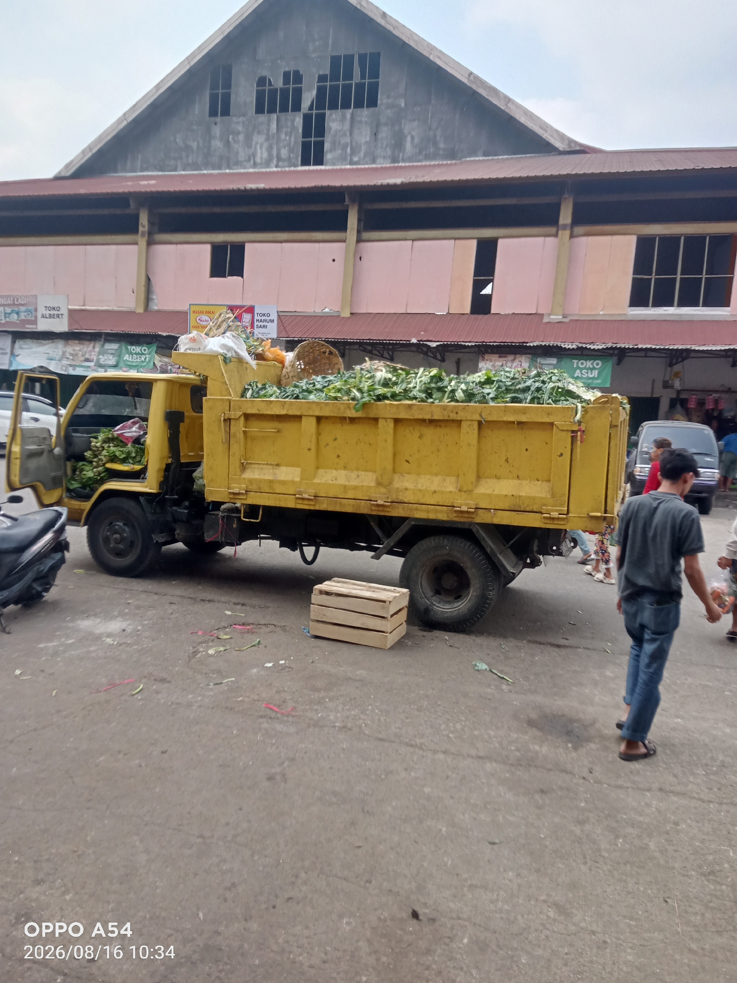 A yellow dump truck loaded with green leafy vegetables is parked on a paved surface in front of a market building. The market building has a large gabled roof and a pink facade with a dark overhang. The truck's driver-side door is open, and some of the vegetables are also visible in the truck's cab. To the right of the truck, a wooden crate is on the ground. Several people are visible in the background, including a man in a dark t-shirt and blue jeans walking away from the camera, and others near a dark minivan. The ground is littered with some scattered green stems and pieces of red plastic. A portion of a motorcycle is visible on the left edge of the image. The scene appears to be an outdoor market in Jambi, Indonesia, with a timestamp indicating August 16, 2026, at 10:34 AM.