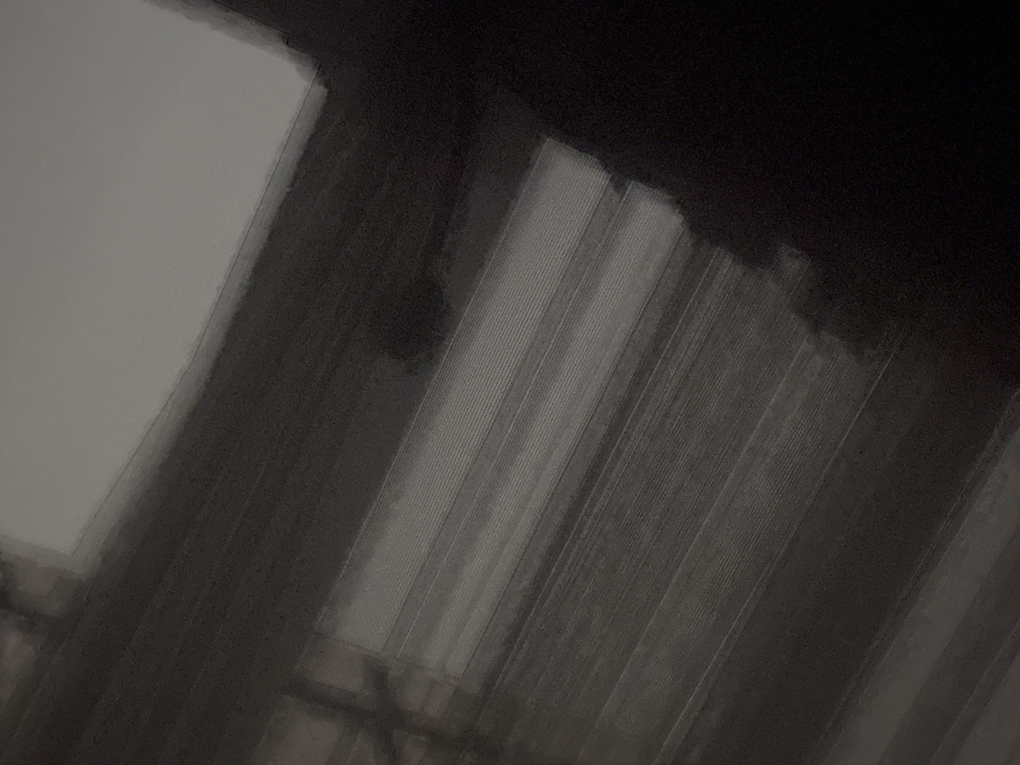 The image shows a low-angle view of an indoor space, with the top portion of the image almost entirely dark. In the center and lower areas, linear patterns suggest the presence of multiple vertical blinds or shades. A lighter, rectangular area appears to be a window or illuminated panel to the left.