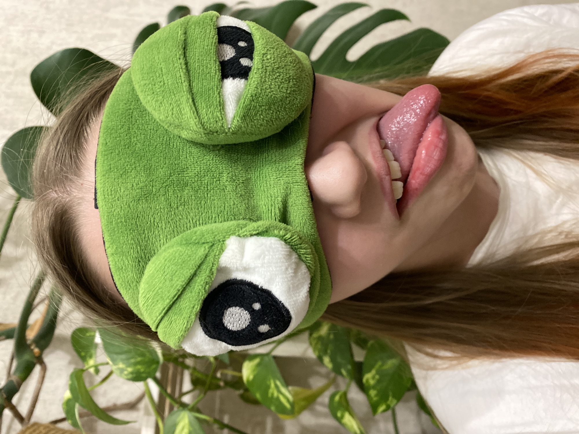 A woman with brown hair is wearing a green frog eye mask and sticking her tongue out. She is lying on a white surface and is surrounded by houseplants. The location is not discernible.