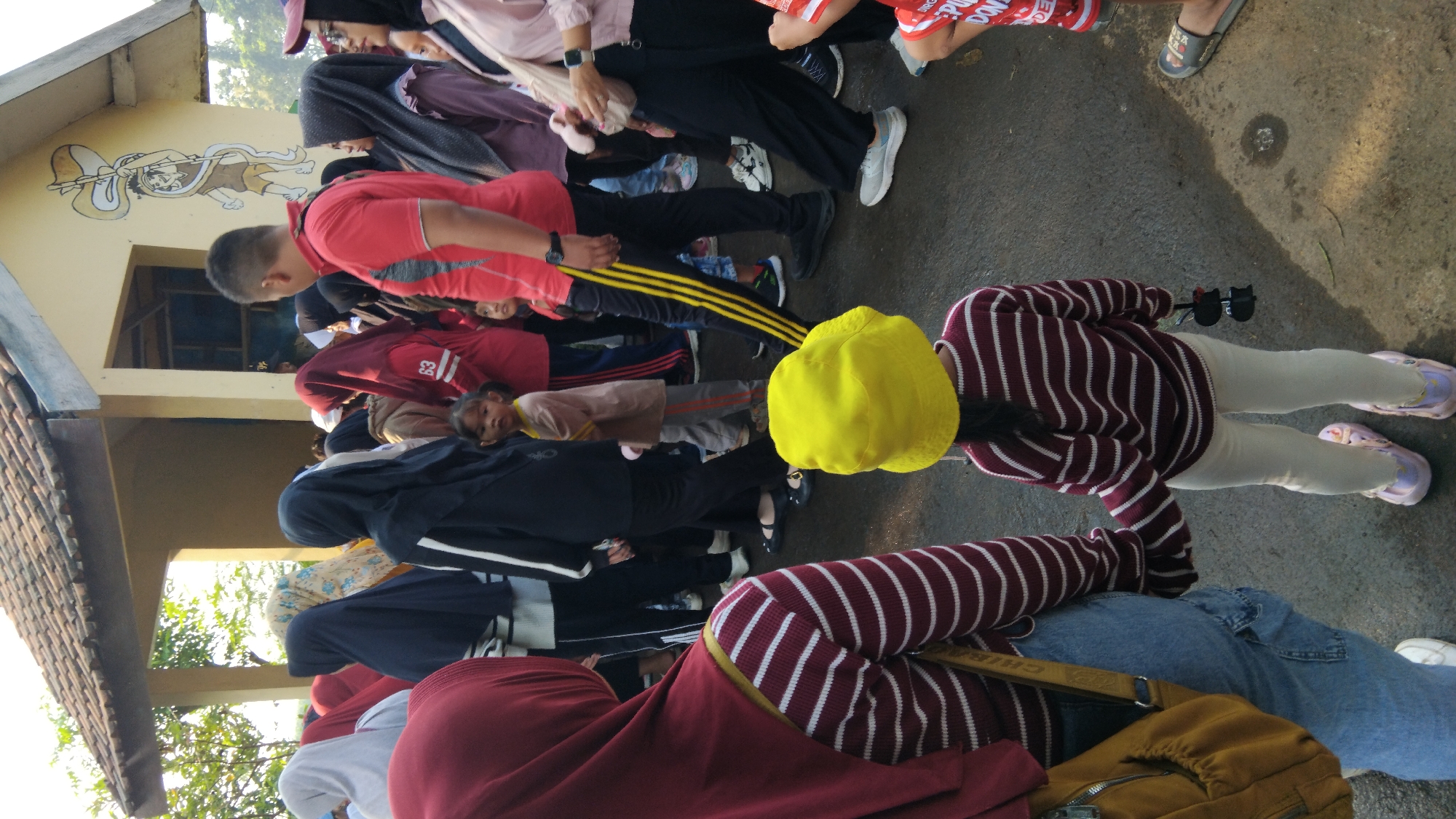 The image depicts a gathering of people outdoors, possibly in the city of Posong, Indonesia, near a building with a tiled roof and yellow walls adorned with a mural of a cartoon character. The focus is on a group of individuals, many wearing headscarves and casual attire. In the foreground, two figures are visible from behind: an adult on the left wearing a maroon headscarf and a burgundy and white striped top, holding hands with a young child on the right. The child is wearing a yellow bucket hat and a matching burgundy and white striped sweater, paired with light-colored pants and pink sandals. The background shows a crowd of people, some appearing to be children, with a variety of clothing and head coverings. The ground is paved and appears wet in places, with shadows suggesting it is daytime.