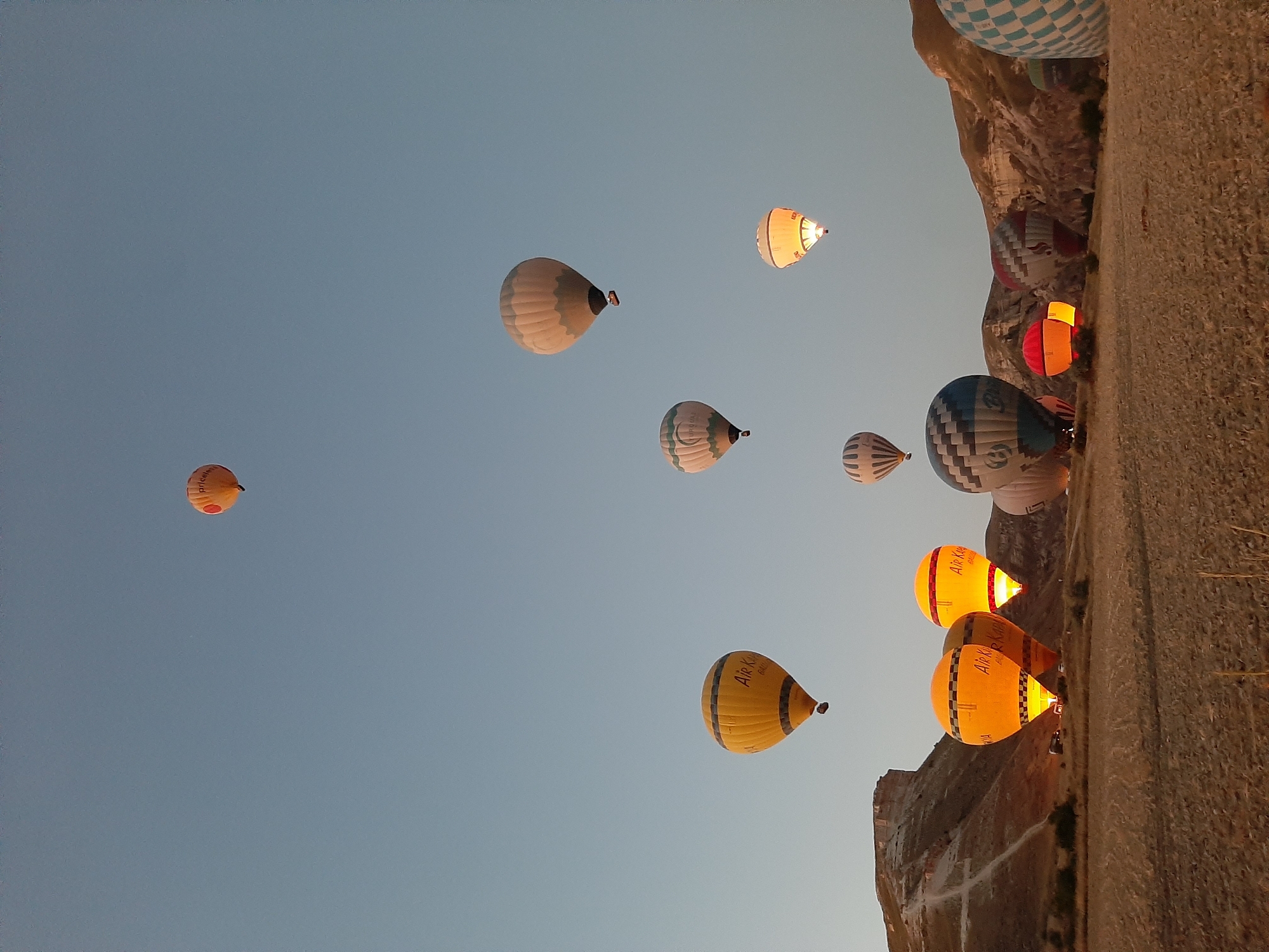 This is a wide-angle, eye-level, outdoor, full shot of several hot air balloons in the sky over a rural landscape, possibly Cappadocia, Turkey, at sunrise. The sky is a clear, light blue. The balloons vary in color and size, with some being brightly lit from within. One balloon in the foreground on the left is yellow with black stripes and the words "Air Kapadokya" written on it. Another balloon to its right is also yellow and lit from within. Further to the right, there are other balloons with various patterns, including a blue and white checkered one on the far right. The ground in the foreground is dry, golden-brown earth, appearing to be a field. In the background, there are rolling hills and rock formations. There are no people visible in the frame.