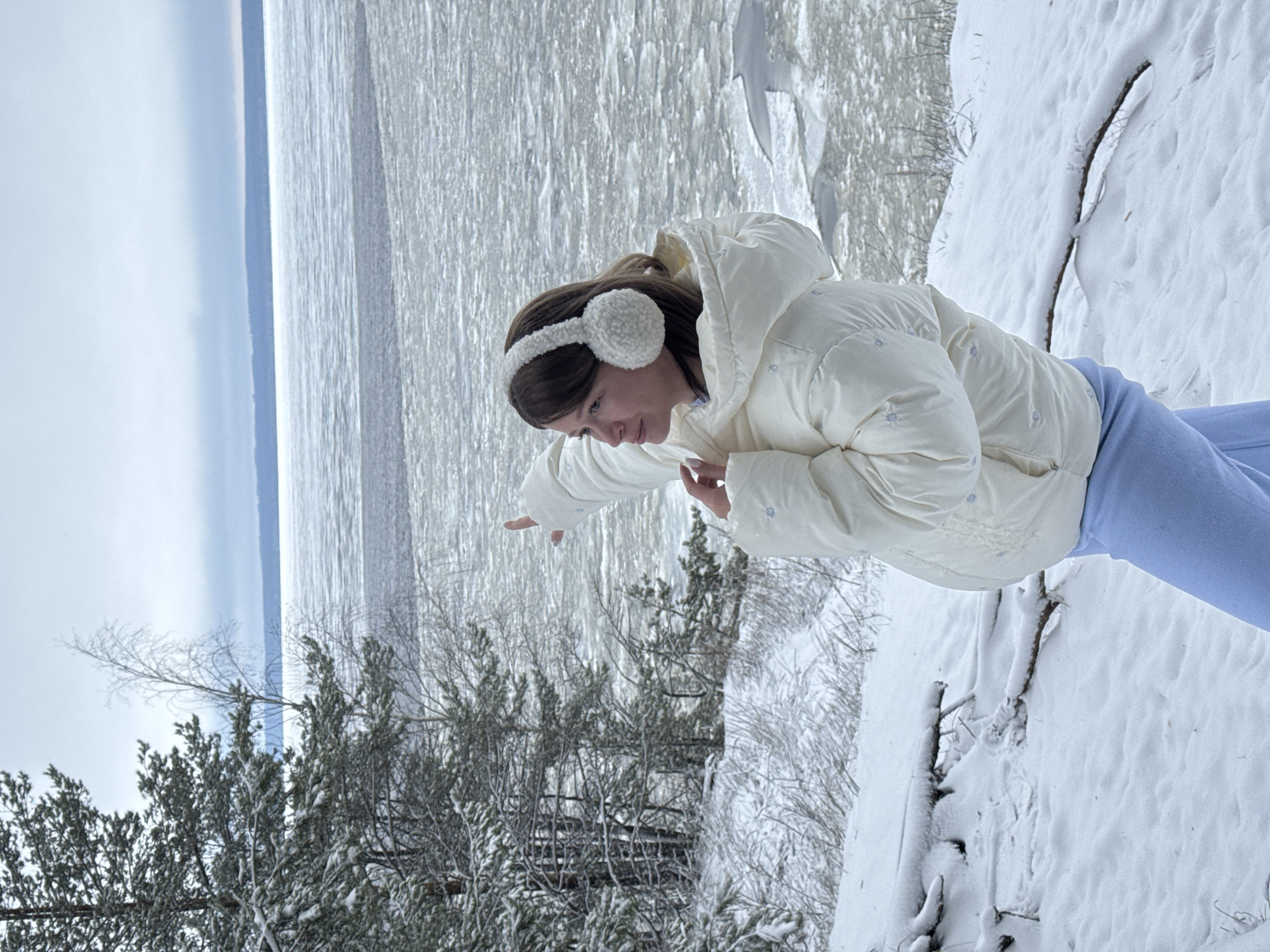 A young woman stands in a snowy landscape. She wears a white puffer jacket with a hood, light blue pants, and white earmuffs. She extends her left arm. The background includes trees and a body of water partly covered in ice. The setting suggests a winter scene in a snowy environment, likely near a lake or river. The location is Noviy, Russia.