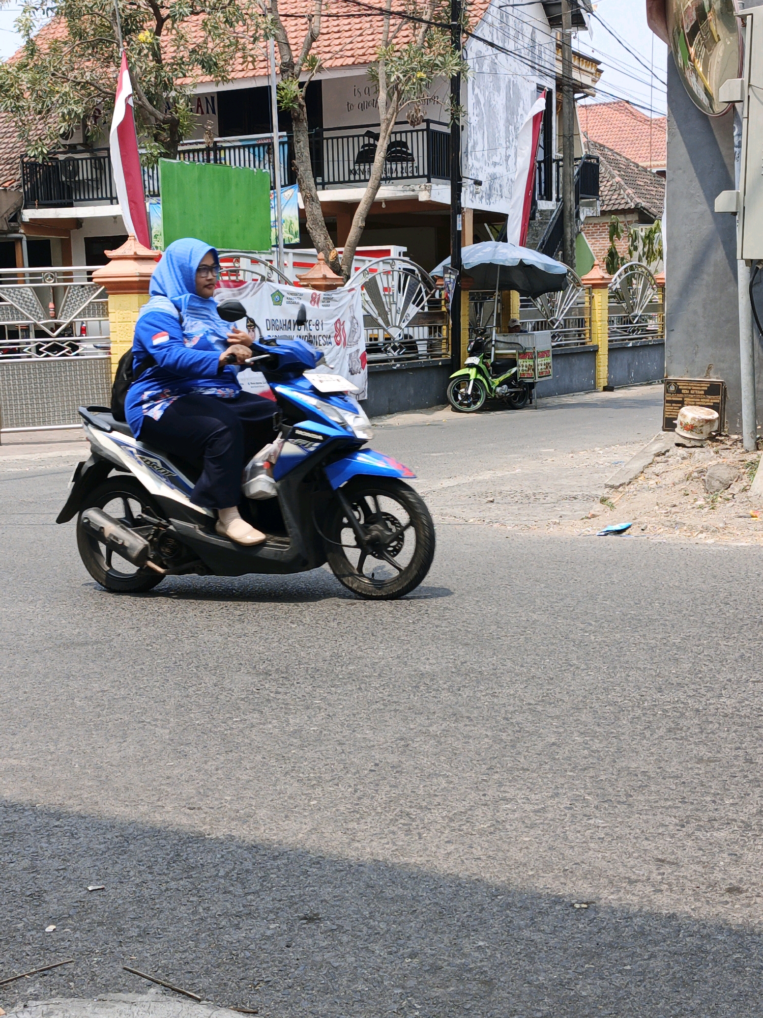 A woman wearing a blue hijab and a blue jacket is riding a blue and white motorcycle on a paved street. She is looking to her left. The street is lined with buildings and fences. A green motorcycle is parked on the side of the street, and a green umbrella is set up next to it. There is a red and white flag hanging on a pole in the background. The scene is likely in Ketegan, Indonesia, as indicated by the signage on the building in the background, which appears to be a school or government building. The weather appears to be sunny.