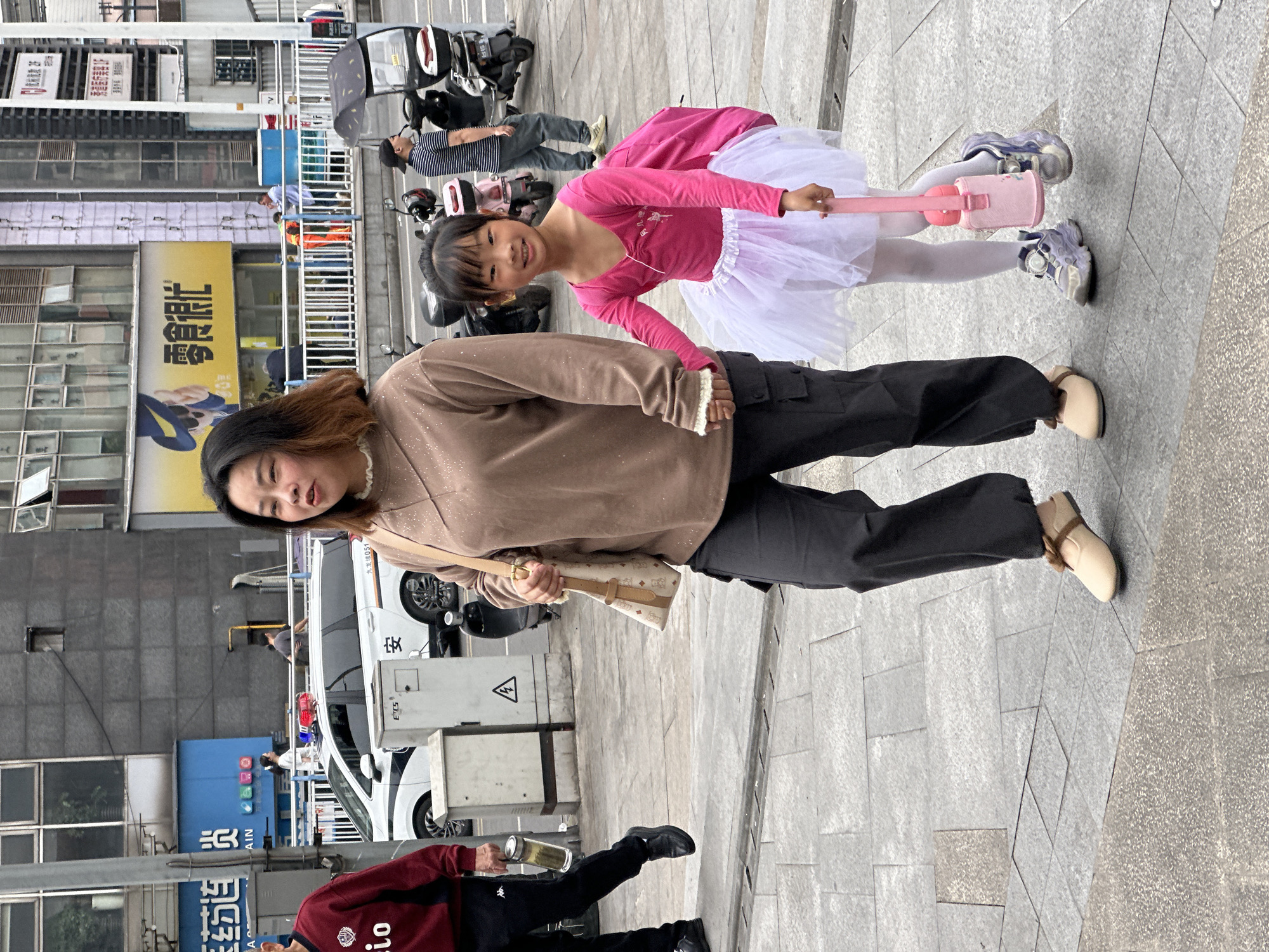 A woman and child stand on a sidewalk. The woman wears a brown sweater, dark pants, and tan flats. She carries a patterned bag over her shoulder. The child wears a pink long-sleeve shirt, a white tutu, and white tights with purple sneakers. The child holds a pink toy. In the background, there are several parked motorcycles, a white van with a triangular warning symbol on the side, a blue sign with Chinese characters, and buildings with advertisements. A man in a red jacket walks by in the foreground. The image appears to be taken in the Jiulongpo District of Chongqing, China, based on the Chinese writing on the signs.