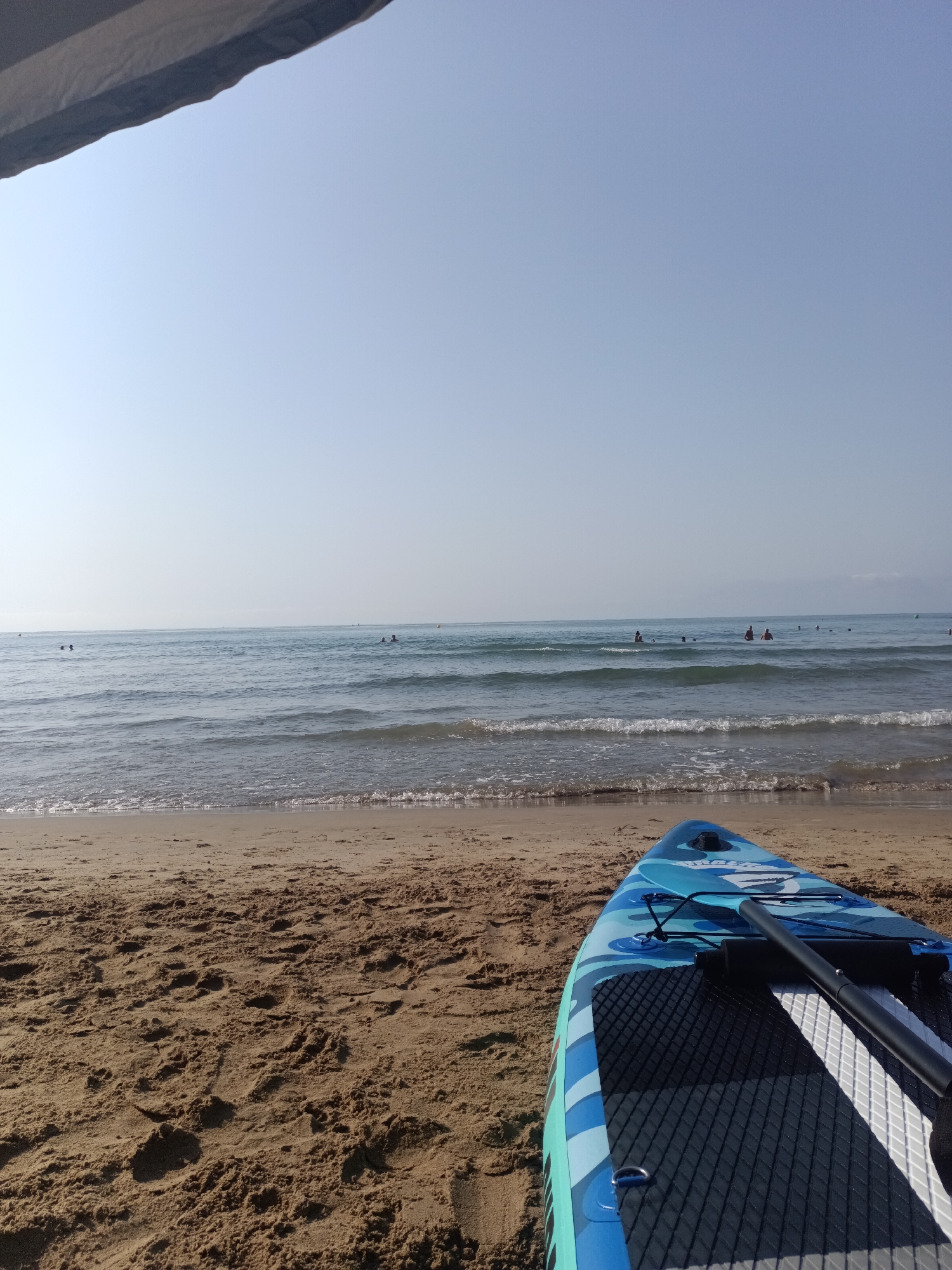 A blue and white patterned stand-up paddleboard rests on the sandy shore of a beach in Orpesa, Spain. The paddleboard, positioned in the foreground facing away from the camera, has a black paddle resting on its surface. The gentle waves of the Mediterranean Sea create a textured surface on the water, with several people swimming and wading in the distance. The sky above is a clear, pale blue with a hint of haziness towards the horizon. Part of a shade structure is visible in the upper left corner of the image.