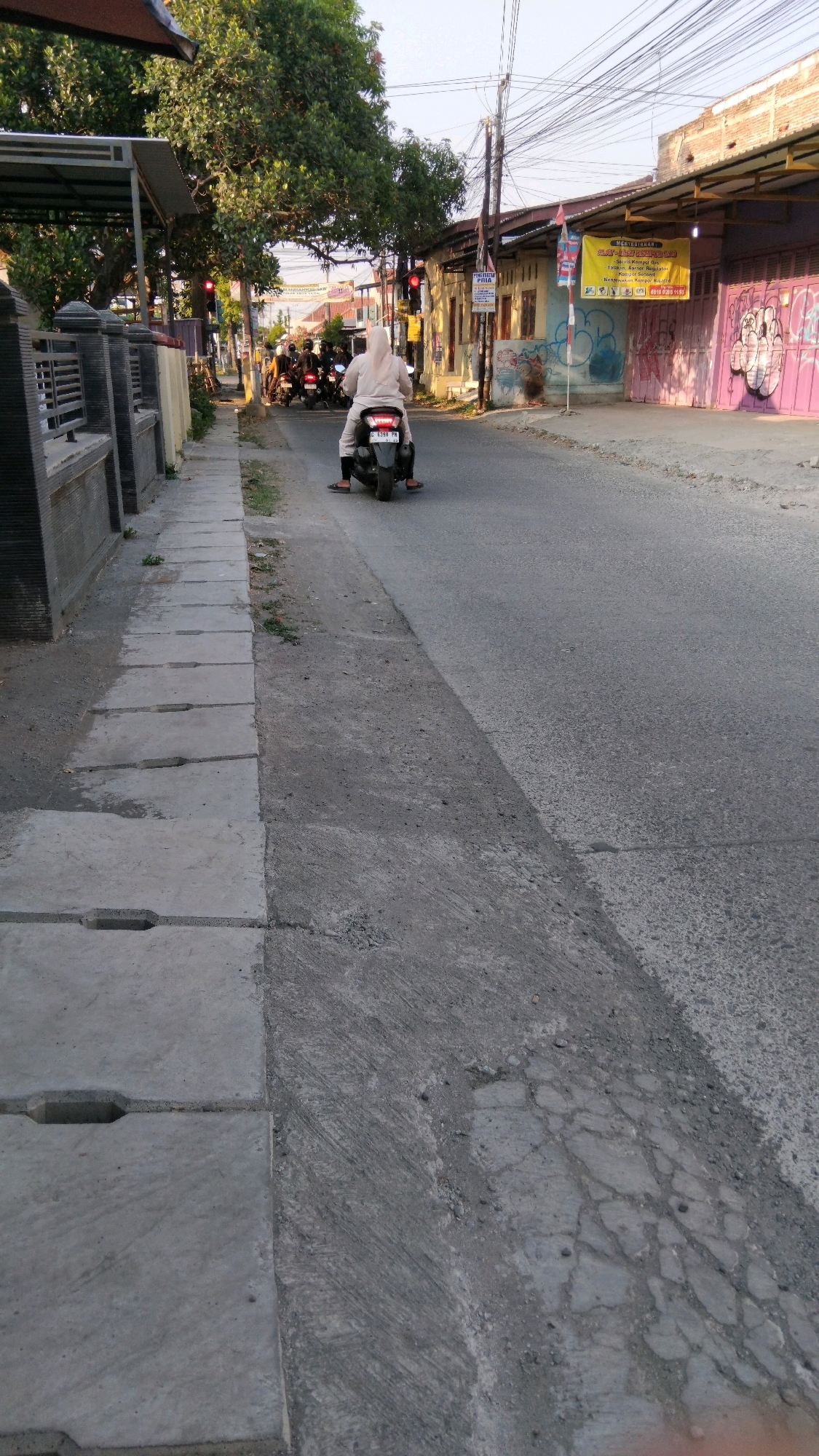 The image shows a street scene in Tegal, Indonesia, likely in a residential or commercial area with moderate traffic. A person wearing a light-colored hijab and clothing is riding a motorcycle away from the camera. The motorcycle's license plate is visible and appears to be from Tegal, displaying the letter 'G' followed by numbers and letters. Several other motorcycles are stopped in the background, suggesting a traffic light or a busy intersection. Buildings line the right side of the street, some with colorful graffiti and signage, including a yellow banner with text and logos. On the left side, a concrete sidewalk with drainage covers runs alongside the road, bordered by a stone fence and vegetation. The sky is clear, indicating daytime. The road surface shows signs of wear and tear with visible cracks and repairs.