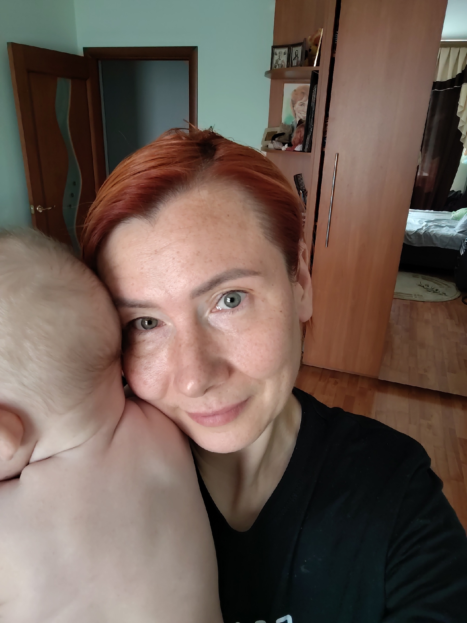 A woman with auburn hair and freckles is taking a selfie while holding a baby. The baby's head is resting on the woman's shoulder, partially obscuring her face. They are indoors, with a wooden door and a large wooden wardrobe visible in the background. A bed with white linens is also partially visible. The scene appears to be in a bedroom in Konakovo, Russia.