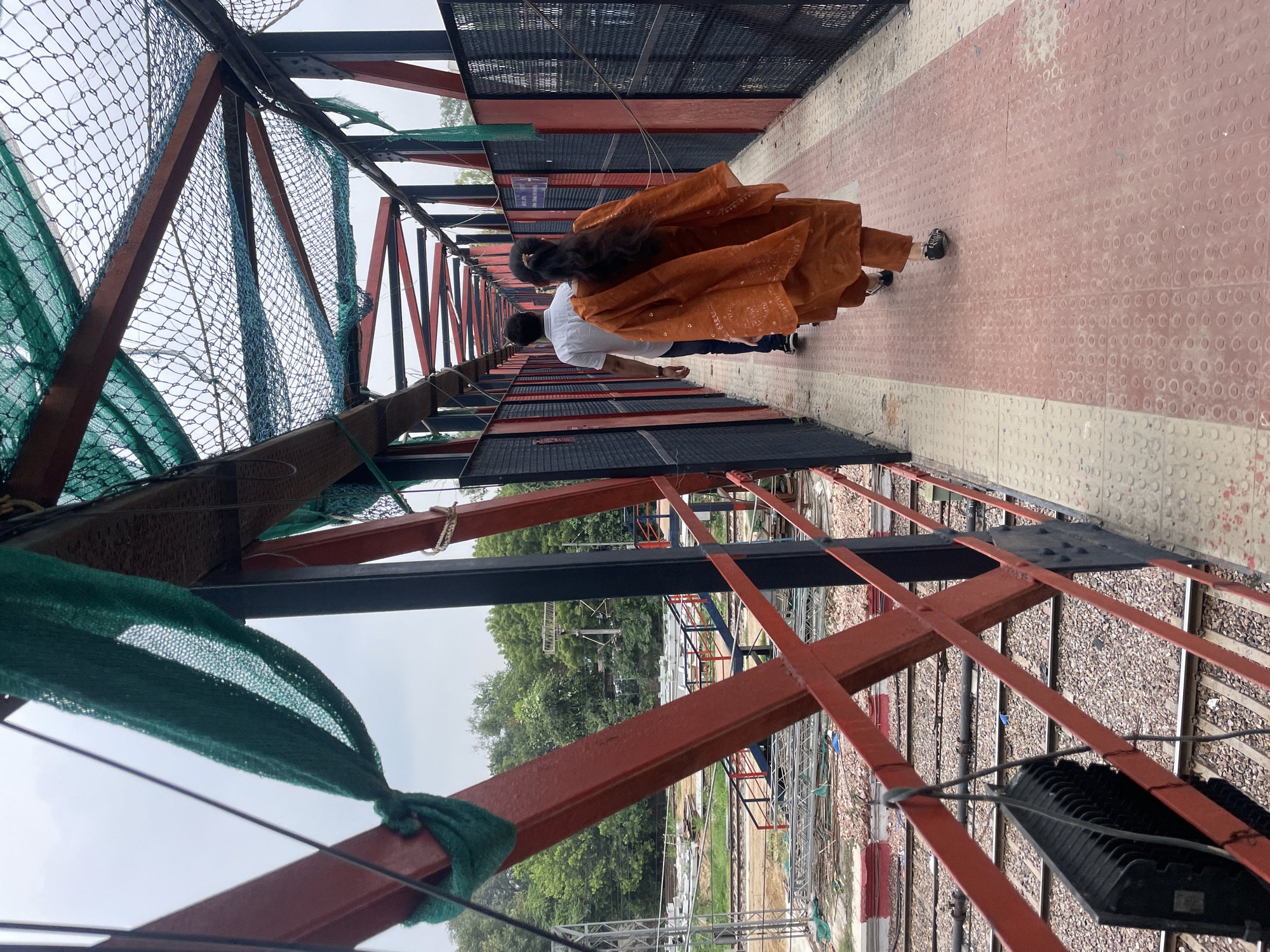 The image captures a pedestrian overpass above railway tracks in New Delhi, India. The overpass features a red metal framework and a pathway made of textured concrete tiles, with a lighter colored section near the railing and a red patterned section in the center. Green netting is suspended above the walkway, presumably as a safety measure. Two individuals are walking away from the camera on the overpass: a man in a white shirt and dark pants, and a woman in an orange traditional Indian outfit, a salwar kameez, with her hair tied back. The surroundings indicate an active railway yard with multiple tracks, gravel, and some construction materials visible below. The sky is overcast, suggesting a cloudy day. The perspective is from the overpass looking forward, with the railway lines receding into the distance.