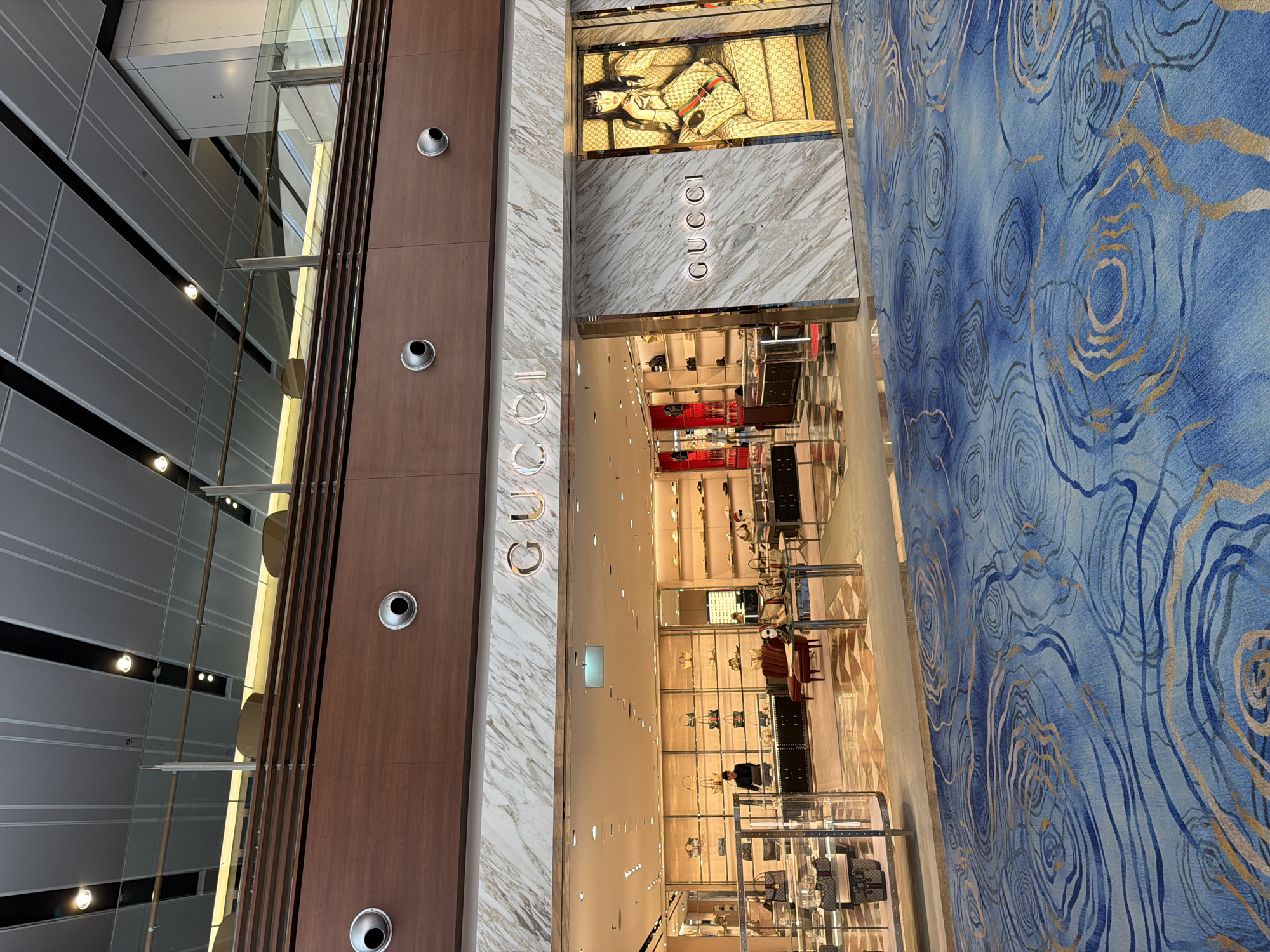 The image shows the exterior of a Gucci store at Singapore Changi Airport's Jewel complex. The store has a marble facade with the "GUCCI" logo illuminated. Inside, merchandise such as handbags and shoes are displayed on shelves. A sales associate is visible behind a counter. The floor in front of the store has a blue carpet with a swirling pattern of gold and blue lines. In the background, the airport's modern architecture with its high ceilings and metallic accents is visible.