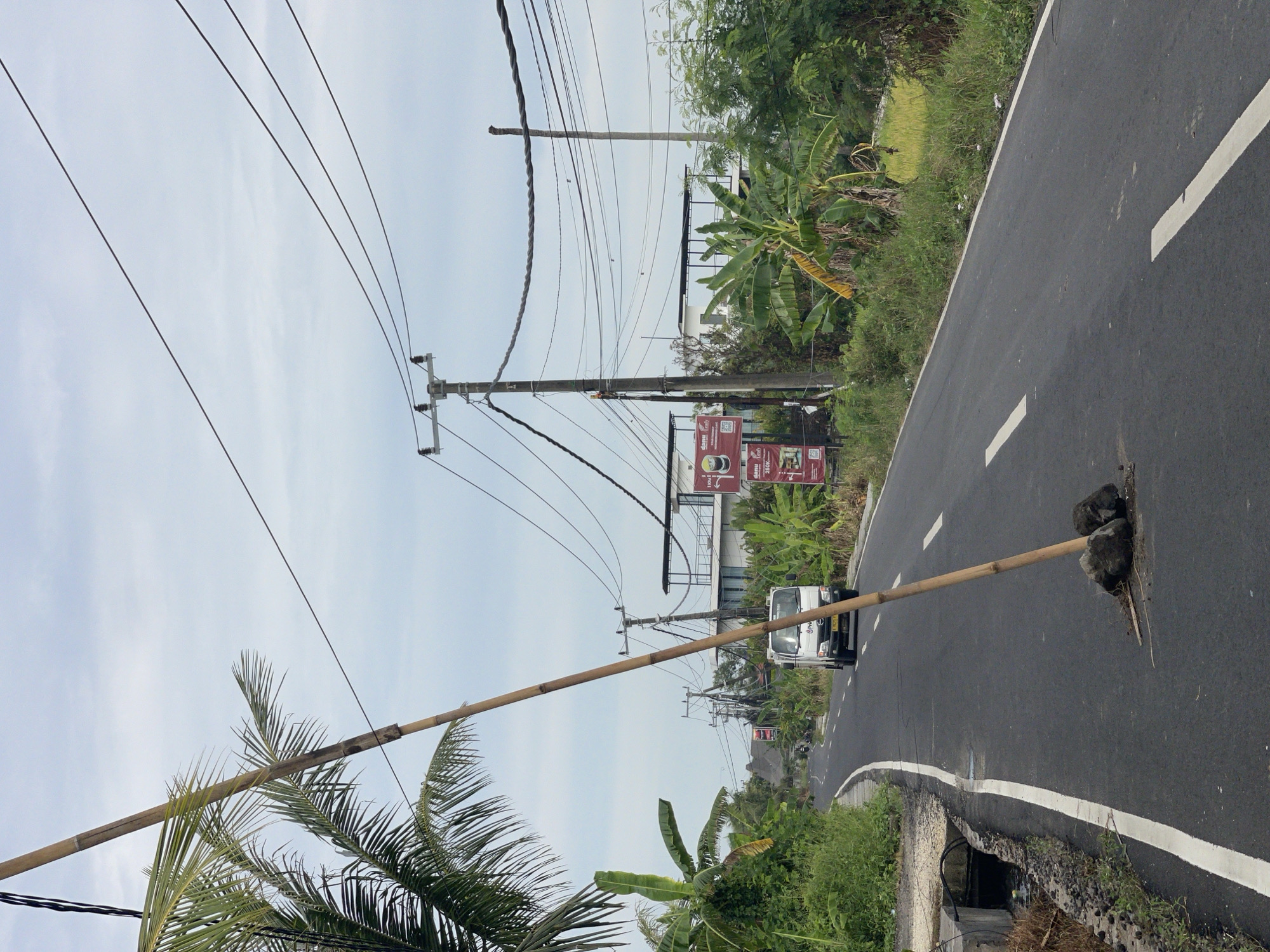 The image depicts a street in Kediri, Indonesia, blocked by a large bamboo pole supported by rocks placed within a pothole. A white utility vehicle is stopped at the improvised barrier. Overhead are power lines, and roadside vegetation including banana trees and palm trees are visible. A sign for a coffee shop is positioned further down the road. The sky is overcast.