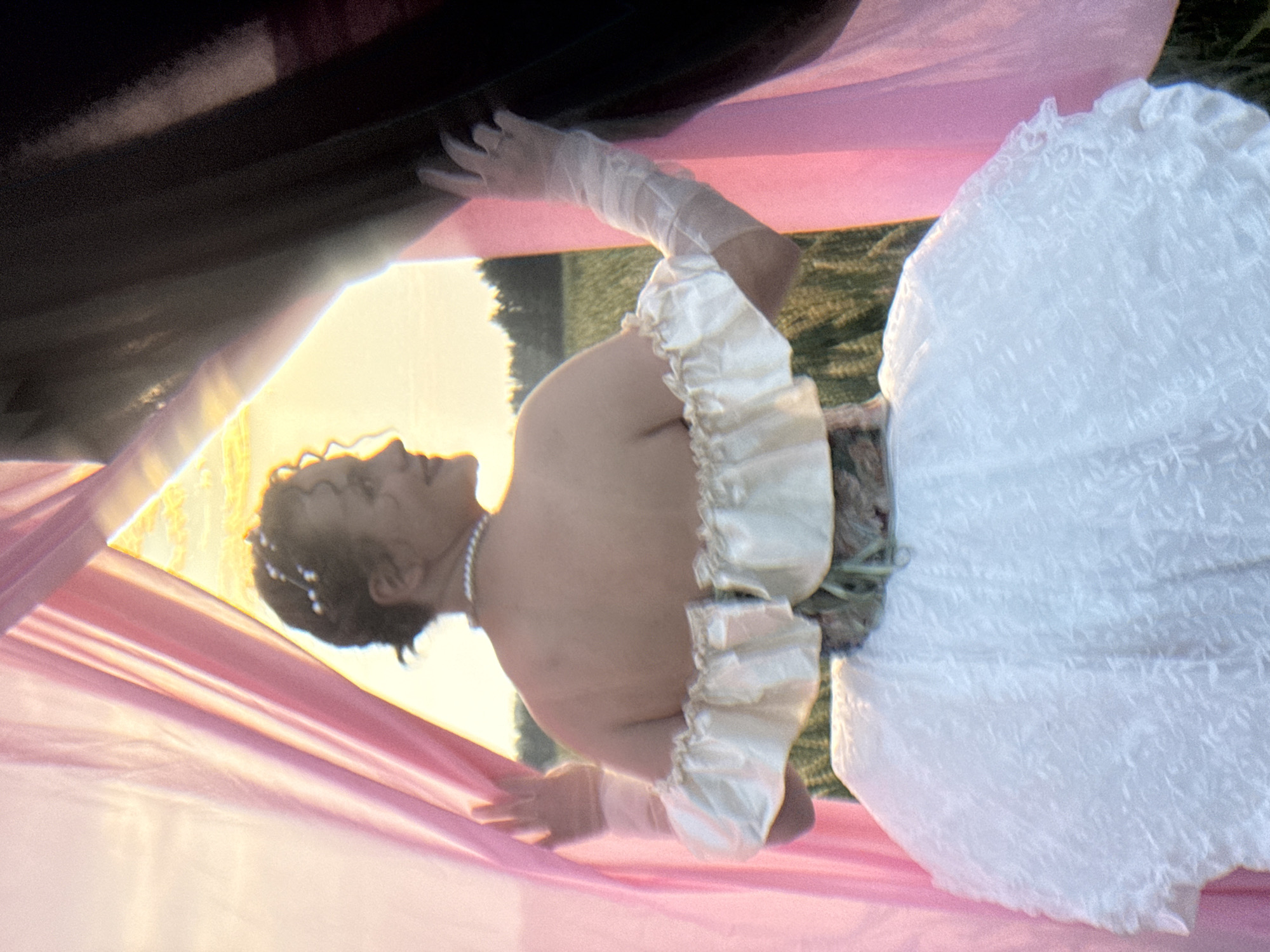 The image depicts a person from behind, wearing a white, off-the-shoulder dress with ruffled sleeves and a voluminous skirt. The person is also wearing sheer gloves and a pearl necklace. They are standing in a field, with a backdrop of trees and a sunset sky. The person is partially obscured by draped pink and black fabric, which appears to be part of a staged setting. The lighting suggests it is late afternoon or early evening. The location is identified as Foki, Russia.
