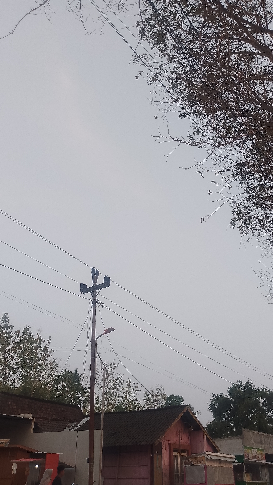 This is a ground-level, outdoor photograph taken during the day in the city of Karanglangu, Indonesia. The scene is dominated by a pale, overcast sky, with a large tree with sparse branches occupying the upper right quadrant of the image. In the middle ground, a utility pole stands tall, supporting electrical wires that stretch across the frame. The pole also has a street lamp attached to it. Below the utility pole, several low-rise buildings with dark roofs are visible, suggesting a residential or commercial area. In the lower portion of the image, a few people are present, though their features are indistinct due to the lighting and distance. One person appears to be wearing a light-colored headscarf, and another is walking. The overall impression is of a quiet, possibly rural or semi-urban environment on a cloudy day.