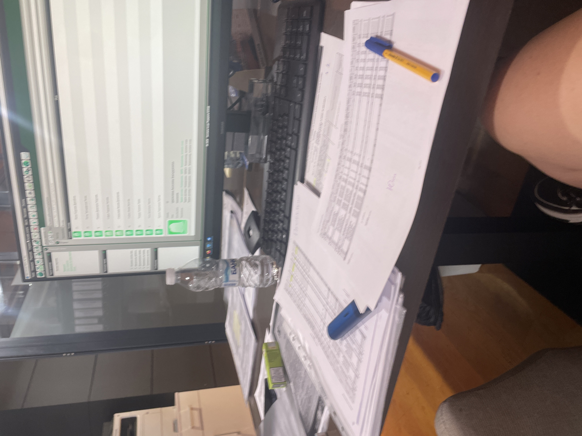 The image depicts a desk in an office environment, likely in Burgas, Bulgaria, given the presence of a bottle of "Bohza" water. The scene is cluttered with papers, some neatly stacked and others spread out. A computer monitor displays a spreadsheet or database with multiple rows and columns of text, along with green icons. A black keyboard is positioned in front of the monitor. To the left, a water bottle and a small green box, possibly containing stationery, are visible. A blue pen lies on top of some documents. In the foreground, part of a person's leg and knee are visible, suggesting the image was taken from their perspective. The overall impression is one of a work-in-progress or an active workspace.