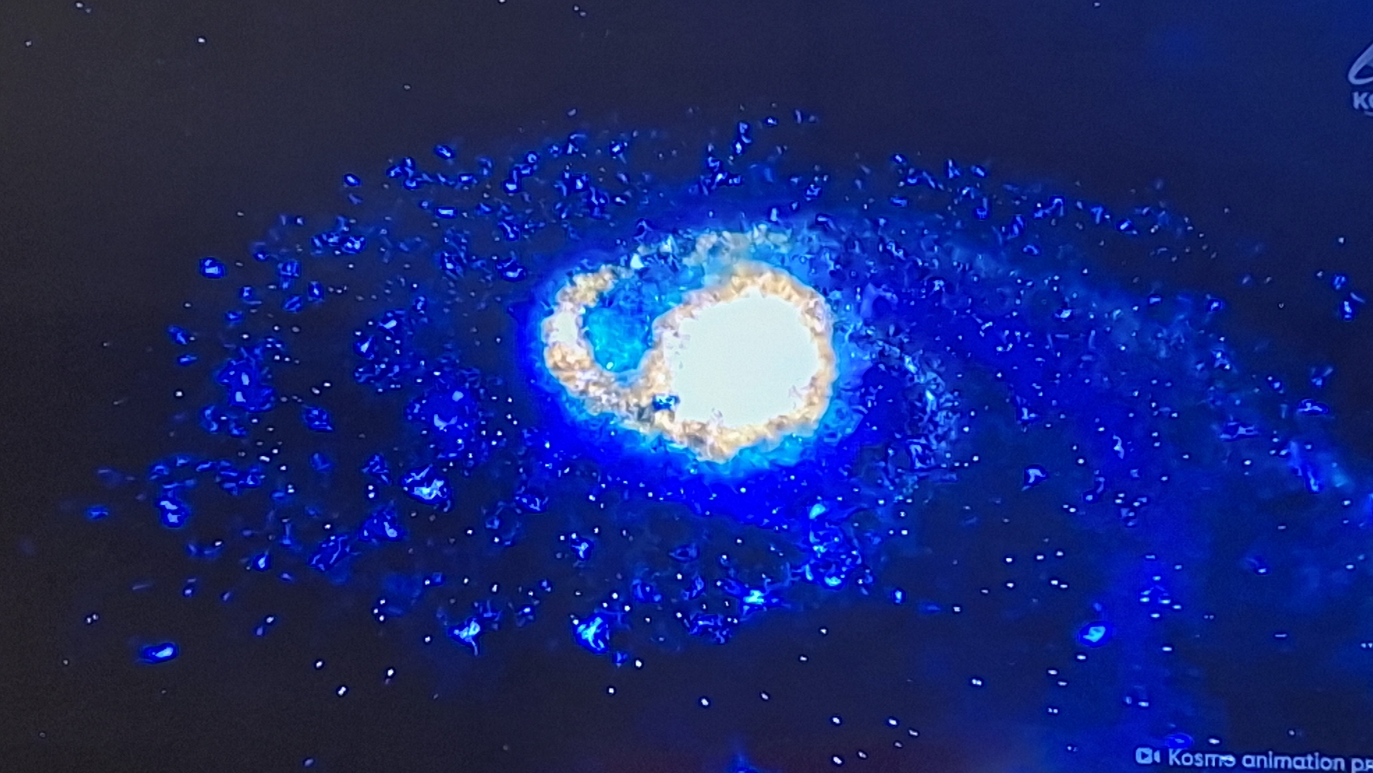 The image shows a digital rendering of a spiral galaxy against a black background. The galaxy has a bright, luminous core, with swirling arms composed of blue and white particles that resemble stars and nebulae. The overall impression is one of cosmic grandeur and energetic motion. There is a logo in the bottom right corner that appears to say "KOSMOS animation pr." This image is a visual representation and does not depict a real-world location in Voinova, Moldova, or any other city.