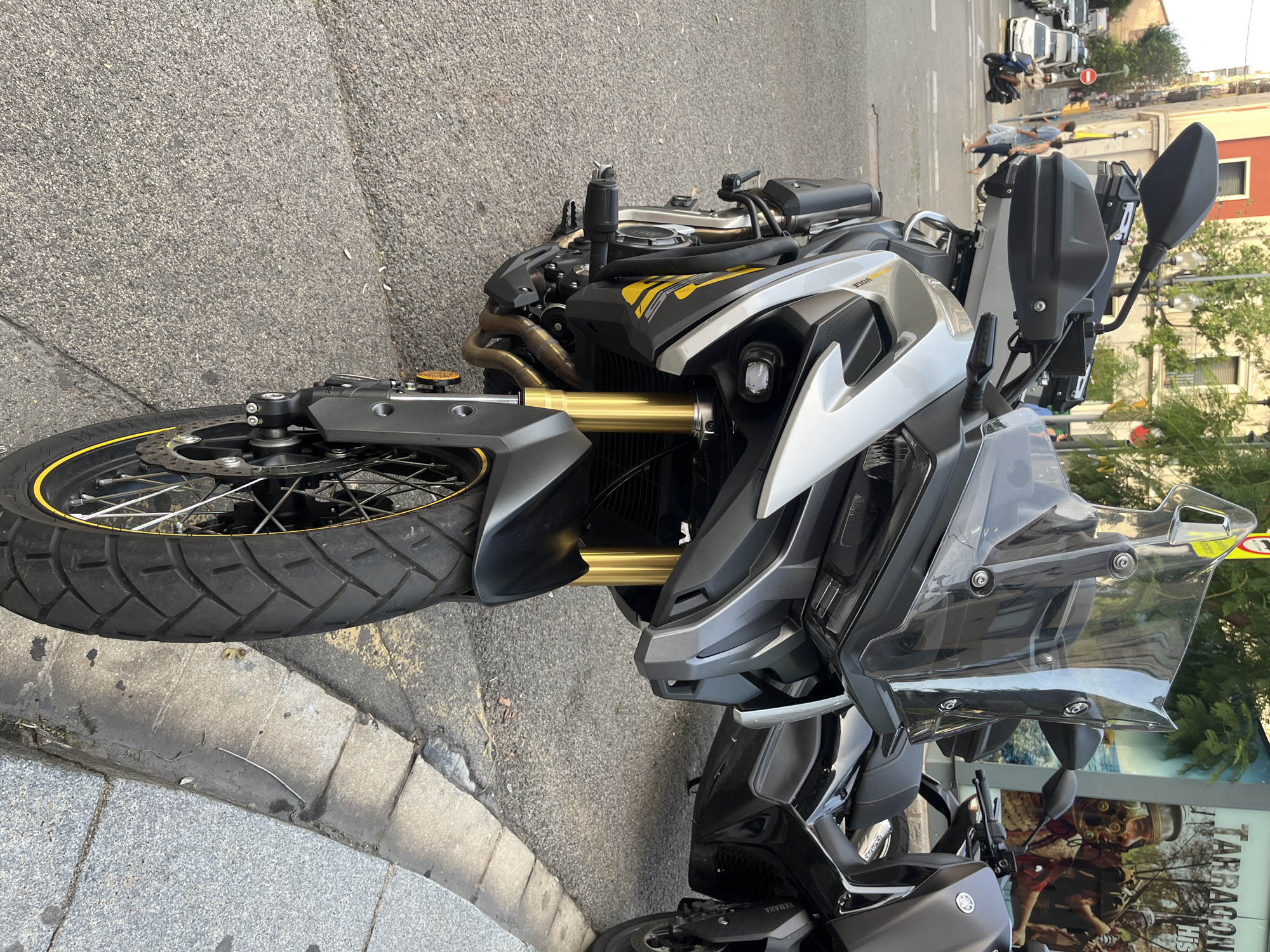 The image depicts a street scene in Tarragona, Spain, with a motorcycle parked on the sidewalk. In the foreground, a silver and black adventure-style motorcycle is prominently displayed. It features gold-colored front suspension forks, a clear windscreen, handguards, and black crash bars. The motorcycle's front wheel, with a yellow accent on the tire, is partially visible. Behind it, another dark-colored motorcycle, possibly a scooter, is parked.
The background reveals a city environment. A building with a reddish-orange facade and balconies stands to the left. Further down the street, several cars are parked, and a few individuals are walking on the sidewalk. One person is riding a scooter. A large poster with images and text, including "TARRAGONA," is affixed to a structure on the right side of the street. The ground is paved with asphalt and cobblestones, with a concrete curb separating the street from the sidewalk. The lighting suggests it is daytime, with sunlight casting shadows.