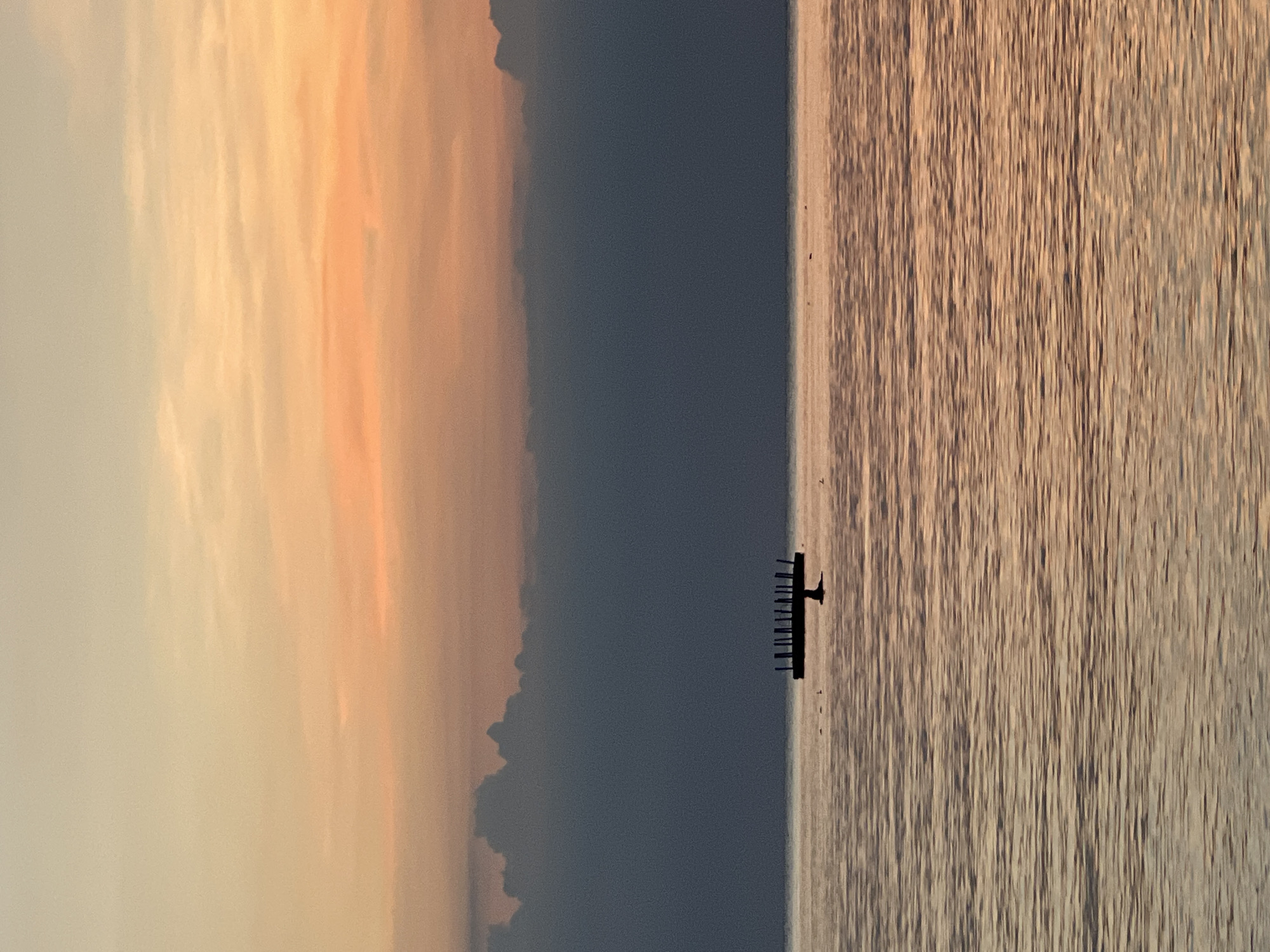 A tranquil scene unfolds at sunset in Ko Pha-ngan District, Thailand, with a calm body of water reflecting the soft hues of the sky. In the distance, a dark, man-made structure, possibly a platform or jetty, rises from the water. Silhouetted against the horizon, it is adorned with several evenly spaced posts, resembling a small, skeletal fence. A few birds are scattered across the water's surface, some near the structure and others further away, adding a touch of life to the serene landscape. The sky above is a gradient of warm colors, transitioning from a pale yellow at the top to a soft orange and pink closer to the horizon, with a band of dark clouds separating the sky from the sea.