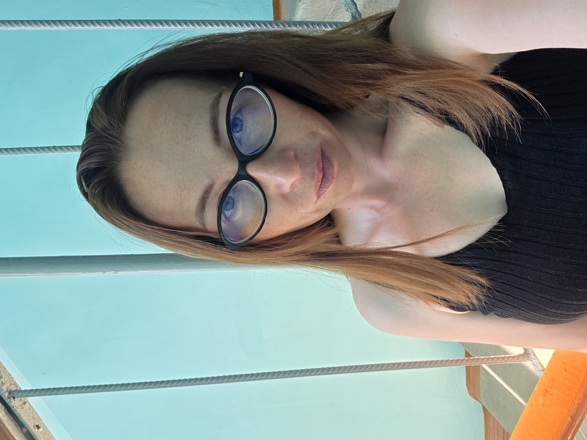 A woman with long, auburn hair and freckles is taking a selfie. She is wearing black-rimmed glasses and a black tank top. She is standing on an outdoor staircase with a teal wall and metal railings behind her. The lighting suggests it is daytime.