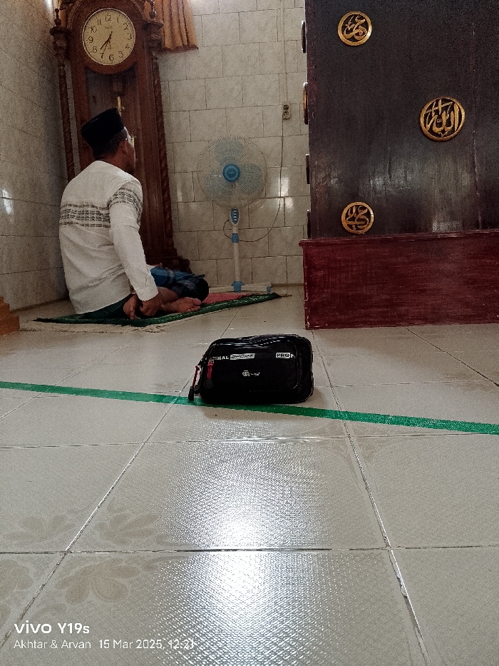 The image shows a man kneeling on a prayer rug in a room with white tiled walls. He is wearing a white shirt and black pants. He is facing away from the camera, with his head bent in prayer. On the floor next to him is a black bag and a green tape line. In the background, there is a wooden cabinet with gold Arabic script on it. A standing fan is also visible, as well as an antique clock on the wall to the left of the image. The room appears to be a mosque or prayer room in Kecamatan Anjatan, Indonesia, based on the provided coordinates. The man appears to be engaged in prayer, with the environment reflecting a religious context. The black bag and green tape suggest there may be a gathering or activity happening within the mosque.