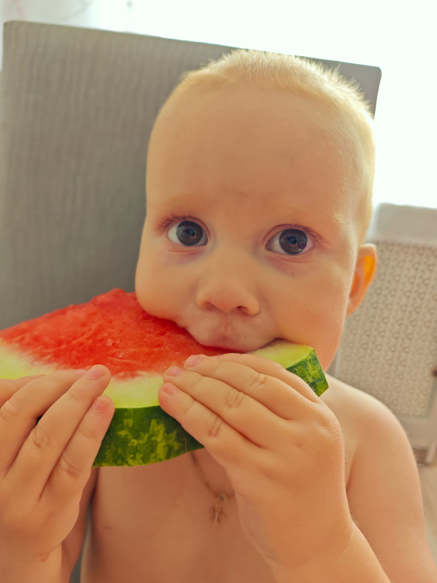 In Markovskii, Russia, a close-up shot captures a young child with blonde hair and wide brown eyes, holding a slice of watermelon to their mouth. The child is wearing a small pendant necklace and their skin has a warm, tanned tone. The background features a blurred gray chair and a white object with a patterned surface. The child's hands are engaged in holding the watermelon, with a visible drop of moisture on their chest.