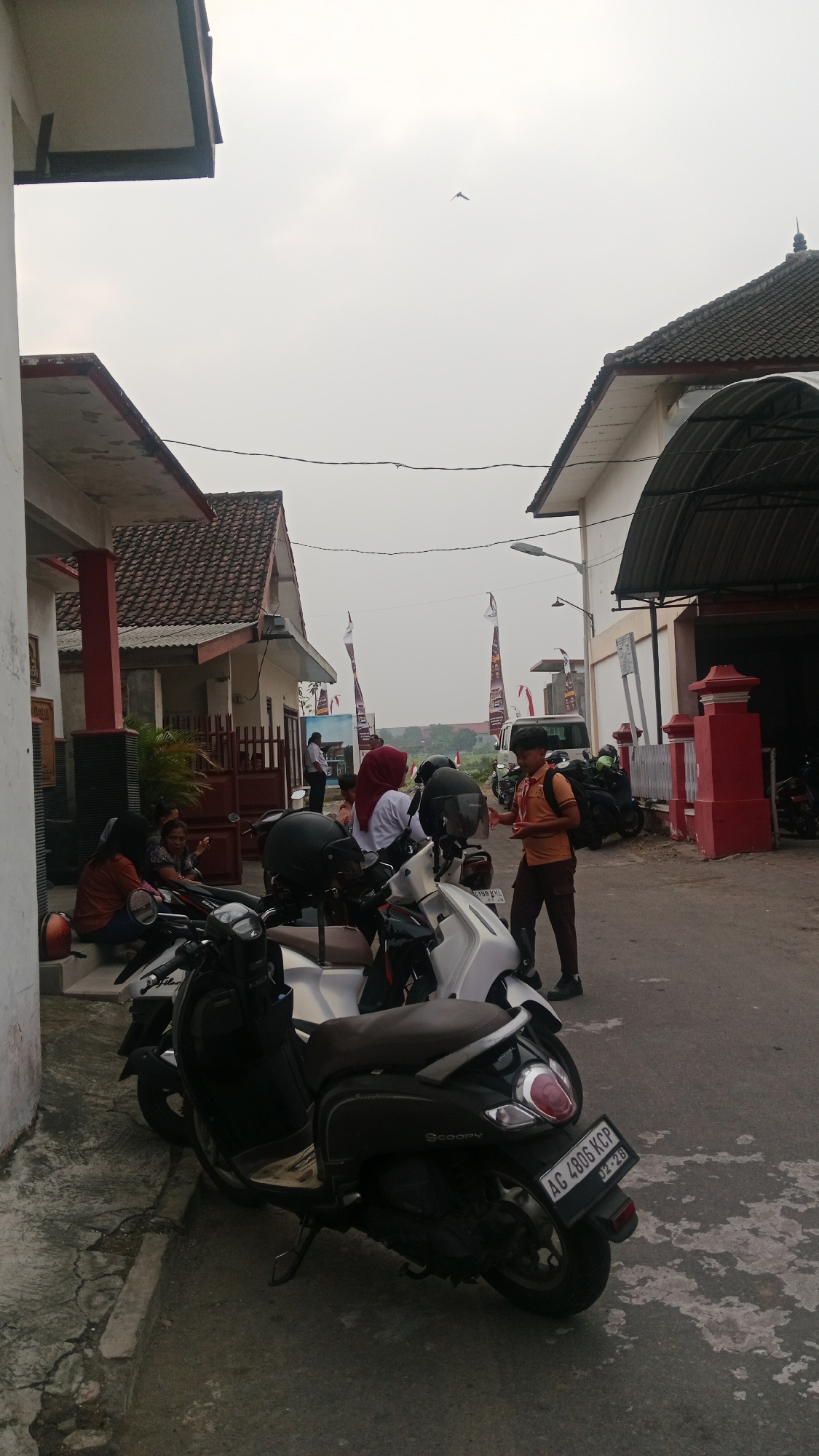 This is a vertical, eye-level, outdoor photograph taken in Blitar, Indonesia. The scene depicts a street area with several parked motorcycles. In the foreground, a dark-colored scooter with a brown seat is prominently displayed. Behind it, a white scooter is visible.
Several individuals are present in the mid-ground. One person, wearing an orange shirt and dark pants, is standing near a white scooter and appears to be interacting with someone. A person in a red hijab and white shirt is seated on a motorcycle. Other people are seated or standing near motorcycles on the left side of the image.
The background features buildings with tiled roofs, some with red accents. Flags with Indonesian text are displayed on poles. A white van is parked in the distance. The sky is overcast, and a small bird is visible in the upper portion of the frame. The ground is paved with concrete.