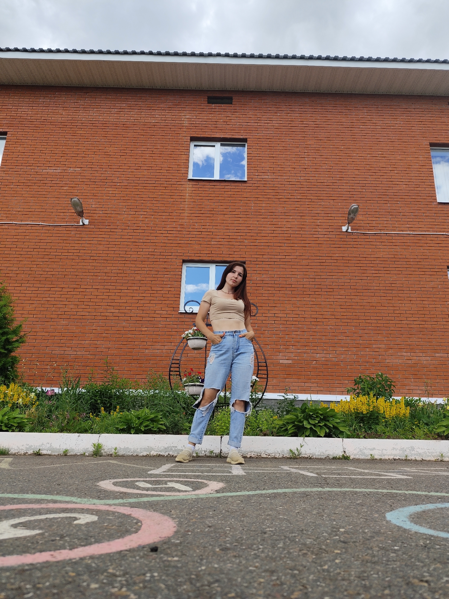 In Фоки, Russia, a young woman stands on an asphalt surface marked with hopscotch grids. She is positioned in front of a two-story brick building, which has two visible windows on its upper level and a roofline with dark gray shingles. The building's facade is made of red brick. Two outdoor light fixtures are mounted on the brick wall. Below the building's windows, there is a flowerbed with various green plants and yellow flowers. The woman is wearing a light-colored cropped t-shirt and light-wash ripped jeans, with her hands in her pockets. She is wearing light-colored sneakers. The sky above is overcast.
