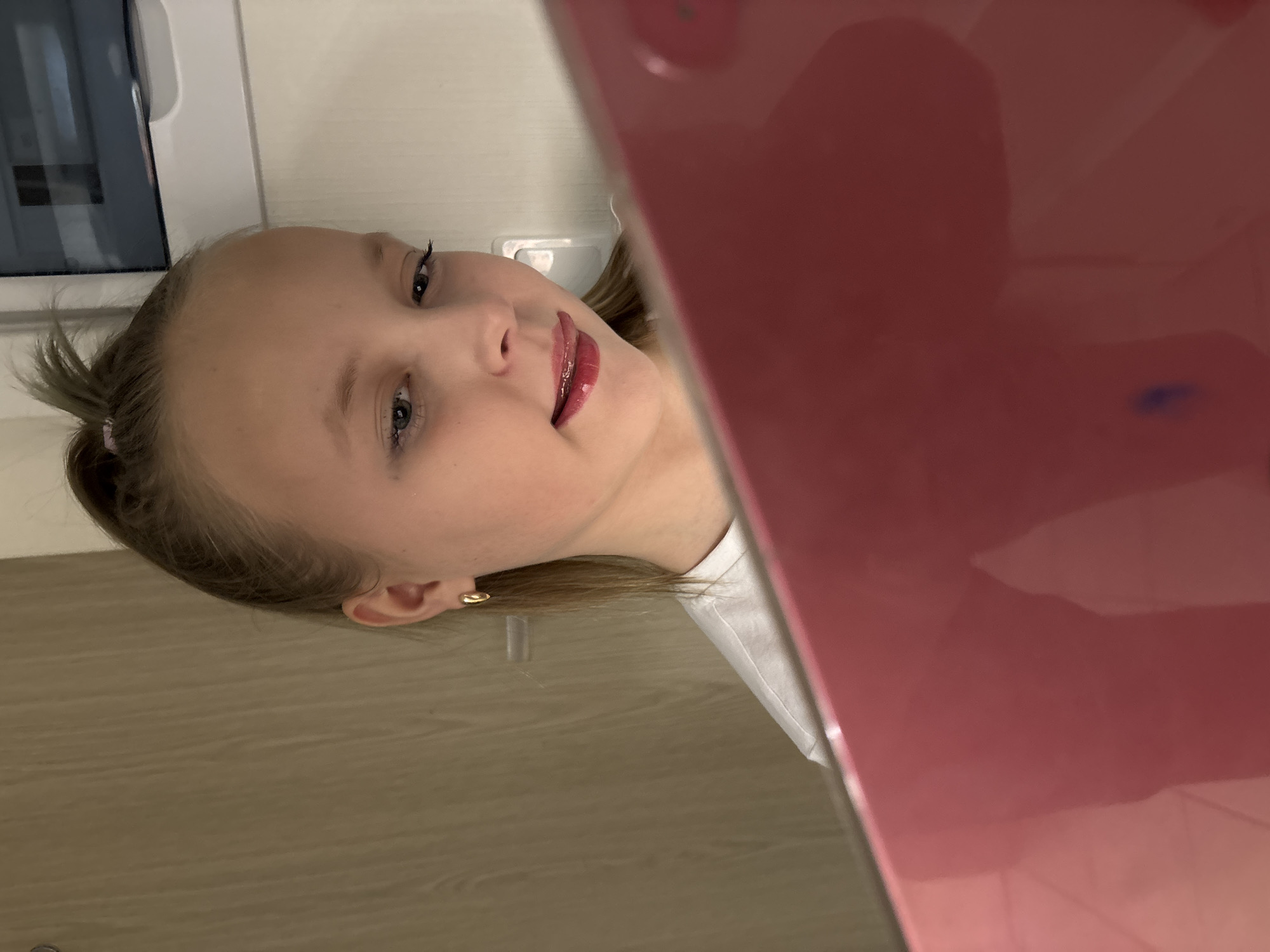 The image depicts a young female in an indoor setting. The individual has light-colored hair, pulled back into a high ponytail secured with a light pink band, and light-colored eyes. She wears small gold-colored hoop earrings in both ears and a white t-shirt. Her lips are coated with a glossy, reddish-pink product, and her tongue is slightly extended between her lips. She is looking downwards towards a bright red or deep pink rectangular object, possibly a tablet or phone, which occupies the lower right foreground and partially obscures her lower face and chest. The background on the left features a vertical surface with a light brown, wood-grain pattern. The background on the right is a plain, light-colored wall, possibly off-white, with a white electrical outlet or fixture visible. Reflections are present on the glossy surface of the red object. The specific location within the city of Izhevsk, Russia, cannot be determined from the visual evidence in the image; however, the scene appears to be a private interior space.