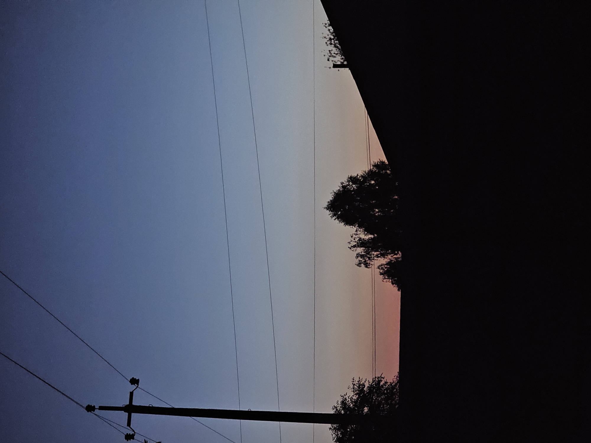 The image depicts a silhouetted rural landscape at dusk in the vicinity of Альняш, Russia. A wooden utility pole with insulators and wires stands prominently on the left, extending upwards into a gradient sky transitioning from deep blue at the top to a pale orange hue near the horizon. Several power lines are stretched horizontally across the frame. In the foreground, the dark silhouettes of trees and a rooftop of a structure obscure the ground. The overall scene suggests an evening in a sparsely populated area, likely a village or countryside, with evidence of infrastructure.
