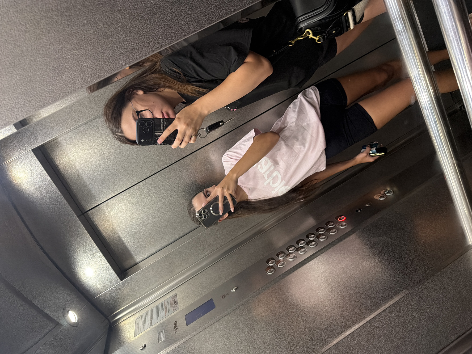Two young women are taking a mirror selfie in an elevator. The woman on the right, wearing a black t-shirt and glasses, is holding a phone with a black case and a keychain. She has a black purse with gold hardware slung over her shoulder. The woman on the left, wearing a pink t-shirt and black shorts, is also holding a phone and appears to be looking at it. The elevator walls are made of brushed metal, and there are visible elevator buttons and a digital display. The lighting is artificial. The image does not provide any information to ascertain the location within Chișinău, Moldova.