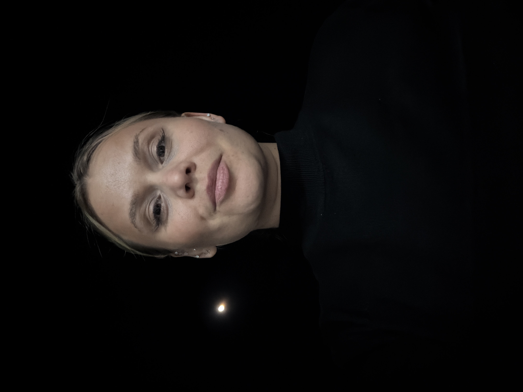 The image presents a close-up, low-angle portrait of a young woman set against a predominantly dark, almost black background. The photo is oriented such that the woman's head is on the left side of the frame, with her body extending towards the right. Her gaze is directed slightly upwards, meeting the camera.
The woman appears to be in her late teens or early twenties, with light skin and blonde hair, which is mostly pulled back or styled away from her face, revealing her features. Her eyes are light-colored, possibly blue or grey, and her expression is neutral with a soft, subtle smile, her lips slightly parted. Small stud earrings are visible in both ears, with what appears to be multiple piercings on her left ear (the one closer to the top of the image in the current orientation). She is dressed in a dark, likely black, high-necked top or sweater, with a ribbed collar visible around her neck.
The background is almost entirely featureless black, indicating a very dark environment, possibly nighttime or a dimly lit indoor space. A singular, distinct detail in the darkness is a small, bright, circular light source located in the lower-middle portion of the frame (if the image were rotated 90 degrees counter-clockwise to an upright position, this light would be in the bottom right). This light has a yellowish-white hue and resembles a distant moon or a focused light in the dark.
The woman's face is brightly illuminated, creating a strong contrast with the dark surroundings, suggesting that a direct light source, such as a camera flash, was used to capture her features. The overall impression is an intimate, low-light selfie or portrait. No text is visible in the image.
