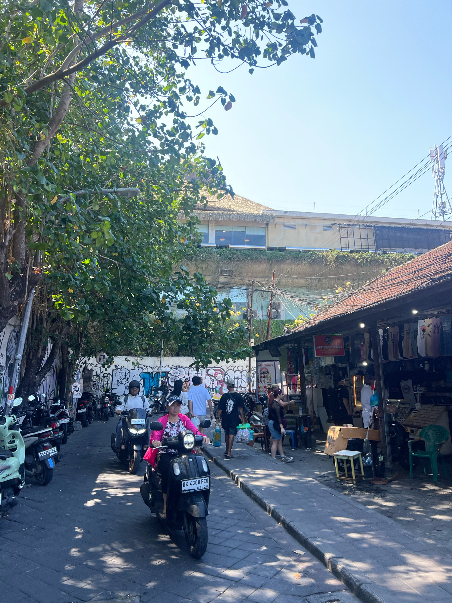 This image depicts a street scene in Kuta, Indonesia, on a bright, sunny day. The foreground is dominated by a narrow, paved street lined with parked motorcycles on the left and a row of market stalls on the right. Several people are present, mostly on motorcycles.
On the left side of the street, a line of scooters and motorcycles are parked along a wall covered in graffiti. A light-colored scooter is parked at the front, followed by several darker-colored scooters.
In the center of the image, two women are riding scooters. The woman in the front is wearing a bright pink patterned top and dark pants and is riding a black scooter. The license plate on her scooter reads "OK 4308 FCQ". Behind her, another person is riding a dark scooter, wearing a white hoodie and a helmet. Further down the street, more motorcycles are parked and a few individuals are walking.
To the right, a market stall is set up under a sloped, tiled roof. The stall displays various clothing items, likely t-shirts, hanging on racks. A red sign with white lettering that says "JIMMY FUCKIN HENDRIX" is visible. Several people are browsing or working at the stall. In front of the stall, there are chairs and boxes.
The background reveals a multi-story building with a thatched roof on the upper level and a lighter-colored facade below. Power lines crisscross the sky above the building. Lush green trees with dense foliage overhang the street from the left, casting shadows on the pavement. The sky is clear and blue. The overall atmosphere suggests a bustling commercial area in a tropical climate.