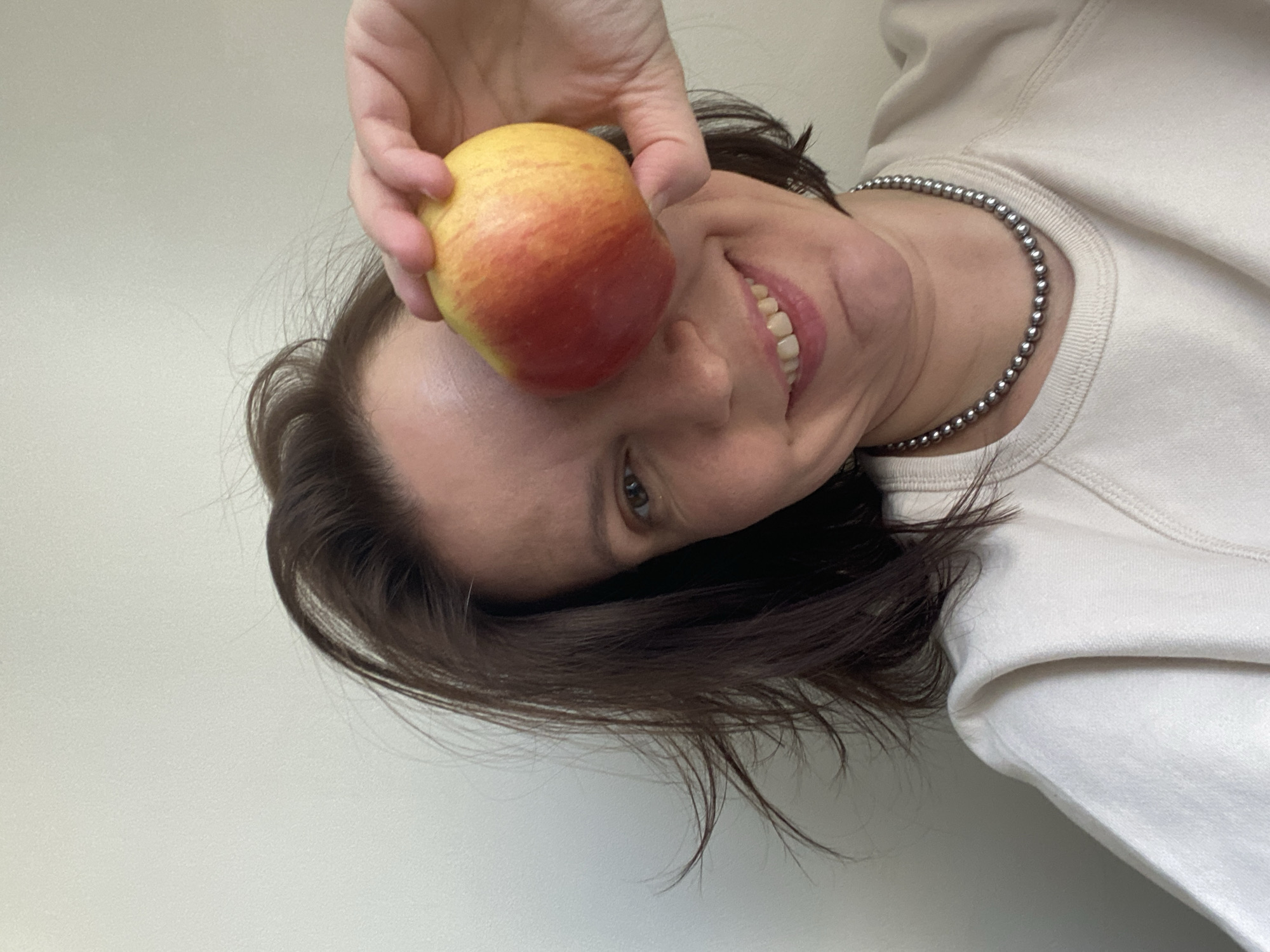 A woman is holding a red and yellow apple near her forehead. She is smiling, and wearing a light-colored sweatshirt and a silver beaded necklace. The background is a plain white wall. The image is rotated counterclockwise. The location cannot be determined from the image.