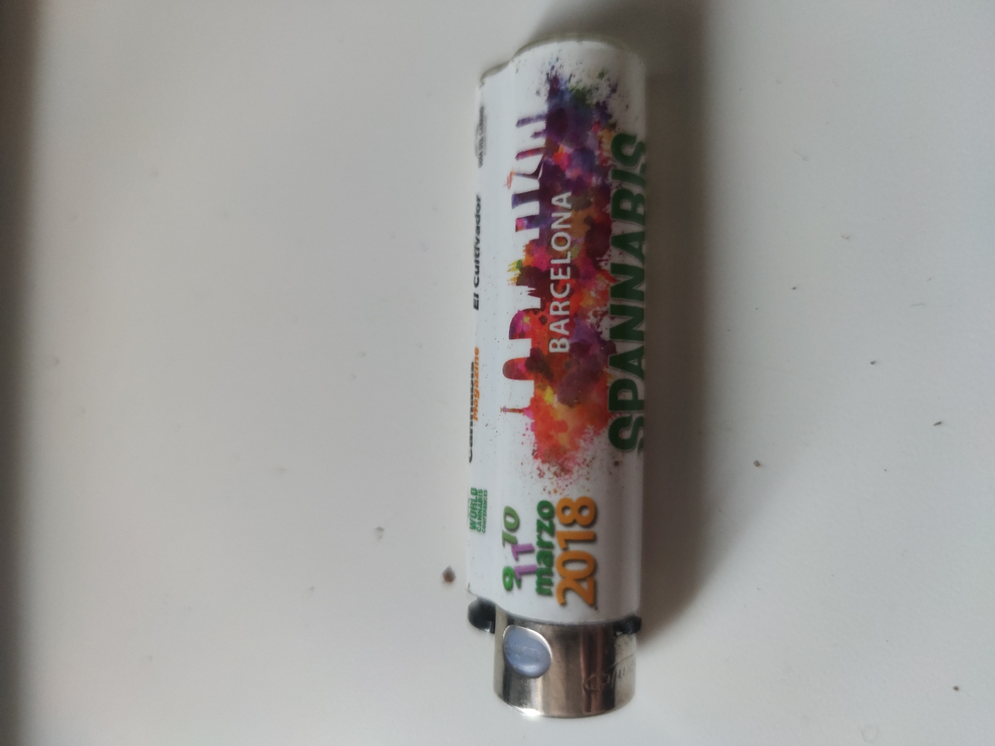 A cylindrical lighter is on a white surface. The lighter has a white body with the text "SPANNABIS" in green letters. Above this text, there is "BARCELONA" in red letters. There are abstract color splashes above the word "BARCELONA". Further up the lighter, there is the phrase "El cultivador" in small, dark letters. Towards the bottom of the lighter, there is "9 10 marzo 2018" in green and orange letters. The base of the lighter is metallic silver. The location is Unknown, Unknown.