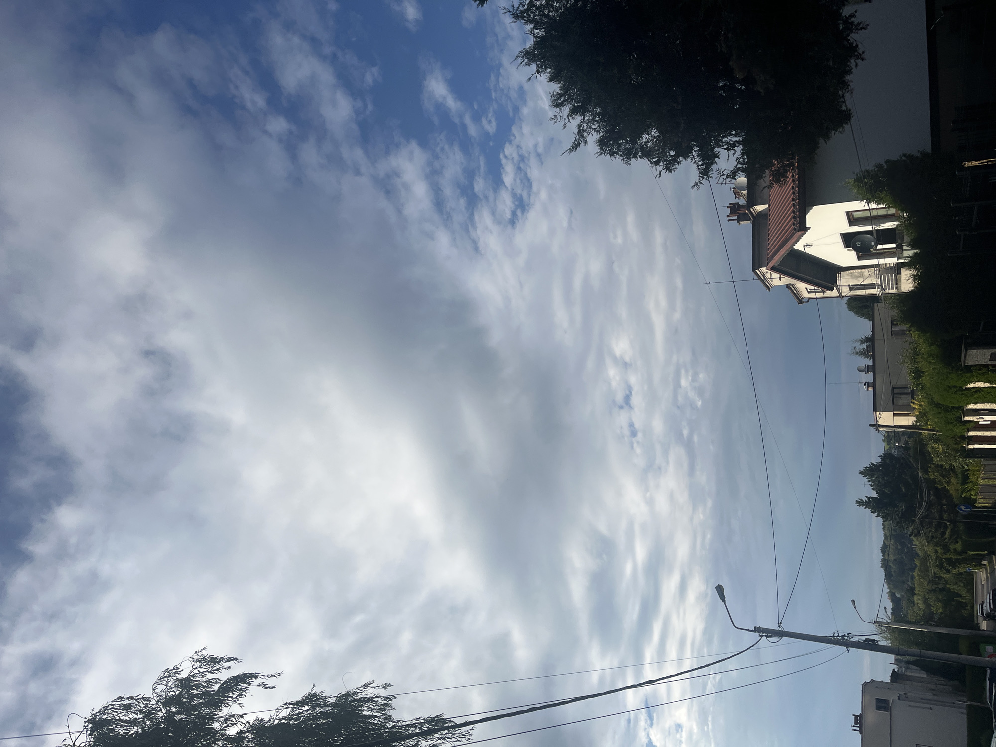 The image shows a street scene with a residential building in the foreground. The building has a white facade, a red tiled roof, and a balcony. There are trees and power lines in front of the building. In the background, there are more residential buildings and a hilly landscape. The sky is partly cloudy with a mix of blue sky and white clouds. The scene appears to be captured during the daytime, possibly in the late afternoon or early evening, given the lighting. The location is in a residential area of a city, with elements of suburban or semi-rural character due to the greenery.