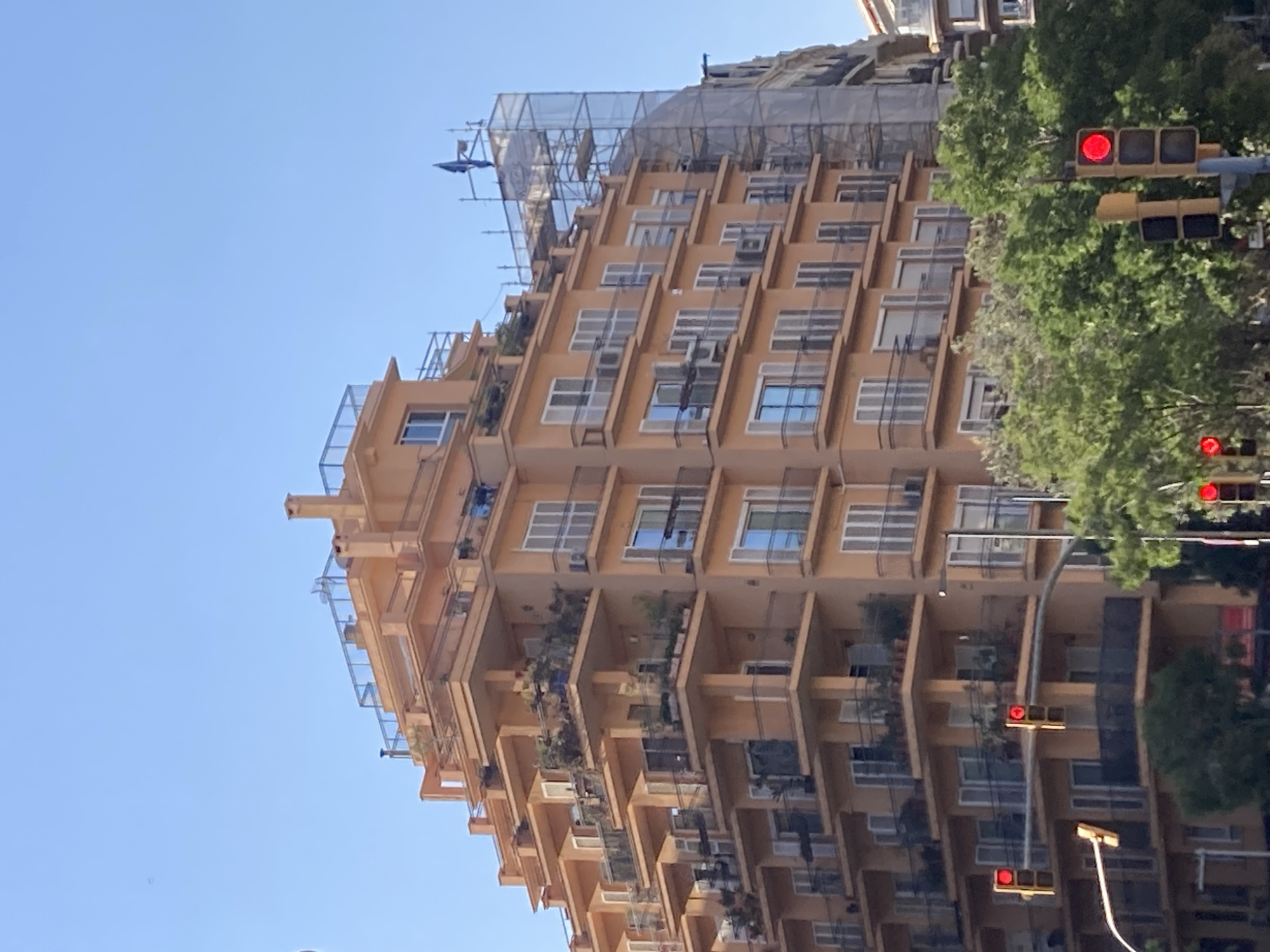 The image shows a multi-story building with a stepped facade, painted in an orange hue. The building features numerous balconies with black railings, many of which are adorned with potted plants. The balconies are set back from the main facade, creating a layered effect. The windows are predominantly white framed. On the right side of the building, scaffolding covered with a white mesh is visible, suggesting ongoing construction or renovation. The sky is clear and blue. In the foreground, traffic lights are visible, with one displaying a red light. Lush green trees are present in front of the building. The architecture and the presence of traffic lights suggest an urban setting, likely a street in Barcelona, Spain.