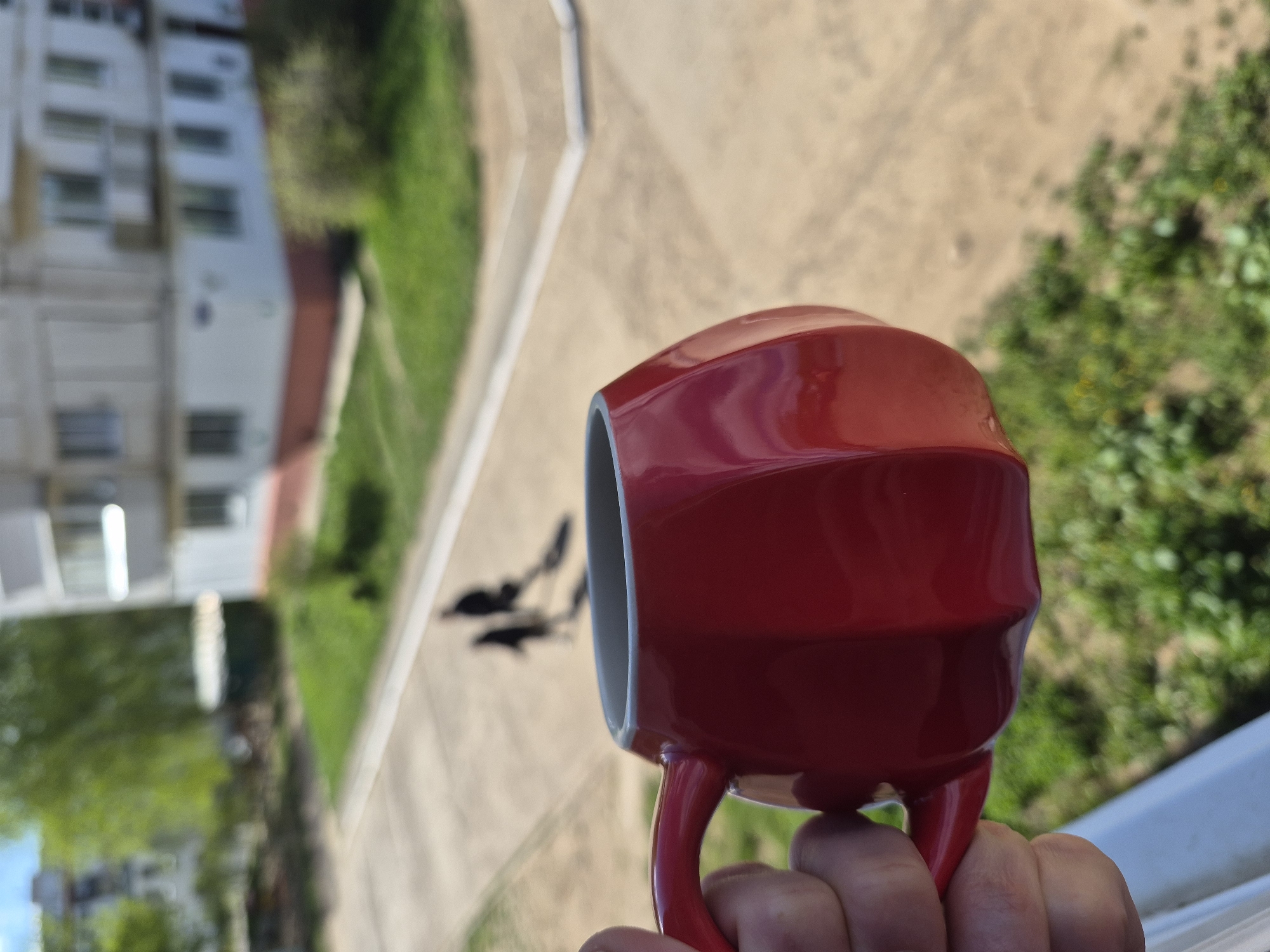 A hand holds a red coffee mug. In the background, slightly blurred, is a courtyard with a sidewalk and grass. A white building is visible on the left side.