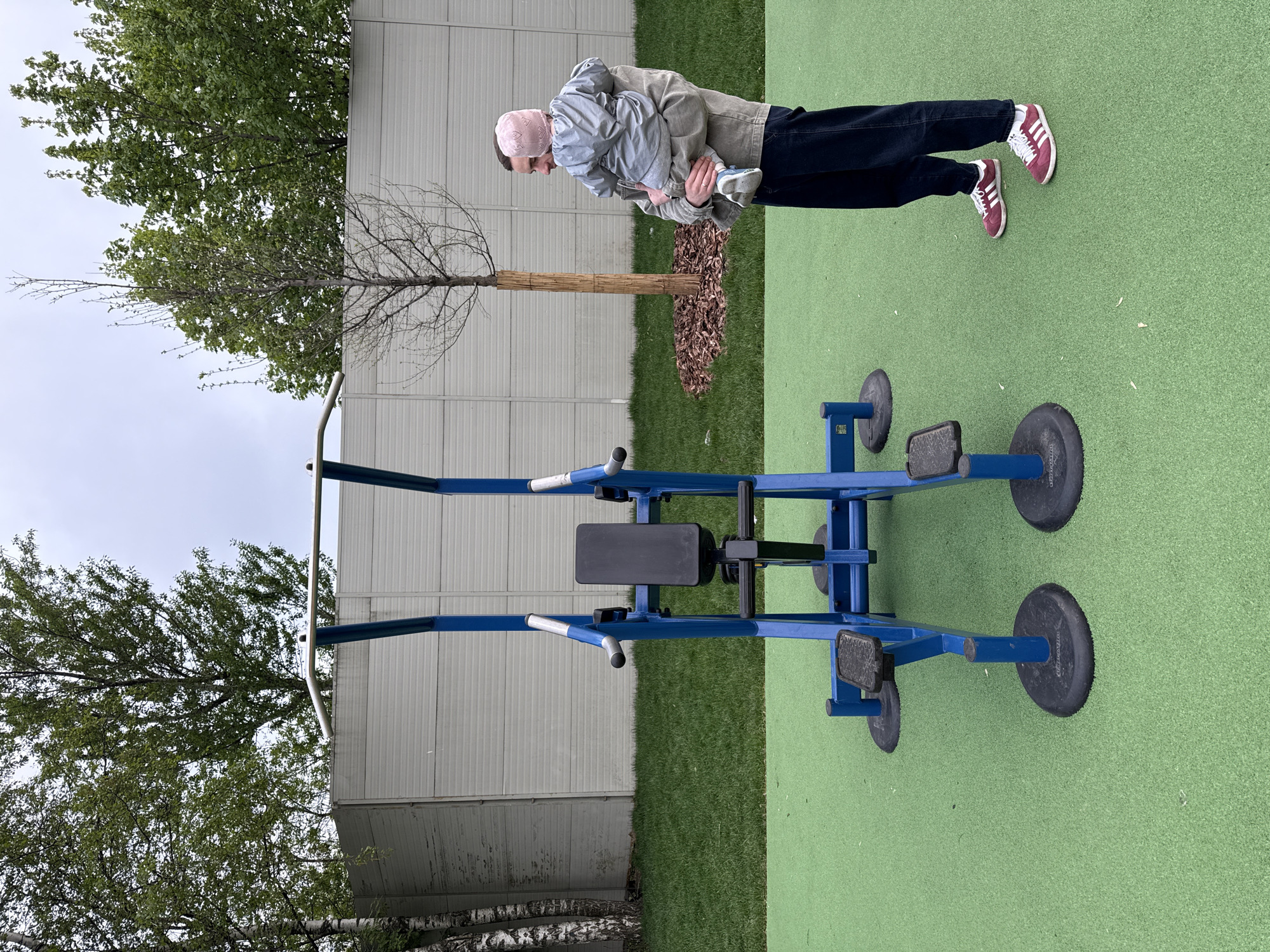 The image captures a scene in Moscow, Russia, showcasing an outdoor fitness area. A man wearing jeans, sneakers, and a light jacket is holding a small child in his arms. The child is wearing a light jacket and a pink hat. They are positioned to the right of a blue lat pulldown machine on a green turf surface. Behind them, there is a tree surrounded by bark mulch, and a gray wall. The sky is overcast.