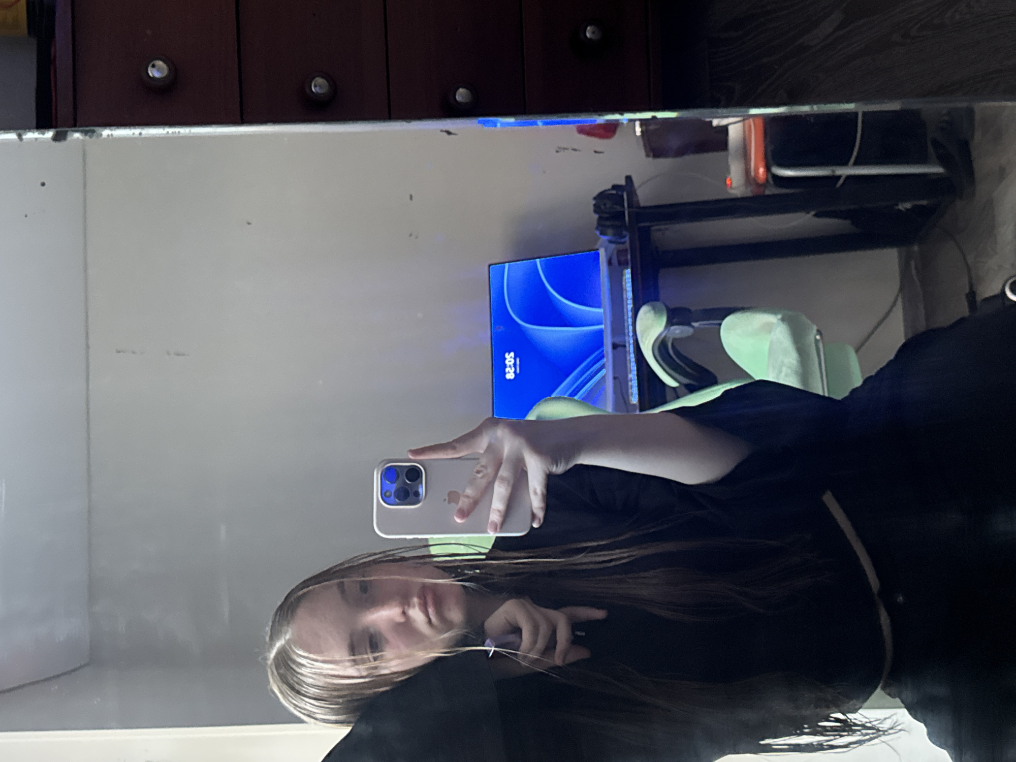 A mirror selfie taken indoors shows a person, likely female, with long brown hair, holding a light-colored smartphone. The individual's face is partially visible, and they appear to be looking into the mirror with a neutral expression.
The reflection reveals a room setup, possibly a bedroom or study area. A monitor displaying a blue abstract wallpaper and the text "50:28" is prominent in the background. Adjacent to the monitor, a light green (or mint) gaming chair is visible. Above the reflection of the room, a dark wooden cabinet or dresser with two silver knobs can be seen. Various wires and cables are noticeable around the computer setup. The mirror itself has some visible smudges or dust. The lighting in the scene appears somewhat dim.
The activity captured is a person taking a self-portrait using a mirror. The time "50:28" on the monitor could indicate a timer, a game time, or possibly a time of day (e.g., 00:50 or 20:50, suggesting evening or late night). The overall setting points to a personal, lived-in space.
The image is contextually located in Perm, Russia.