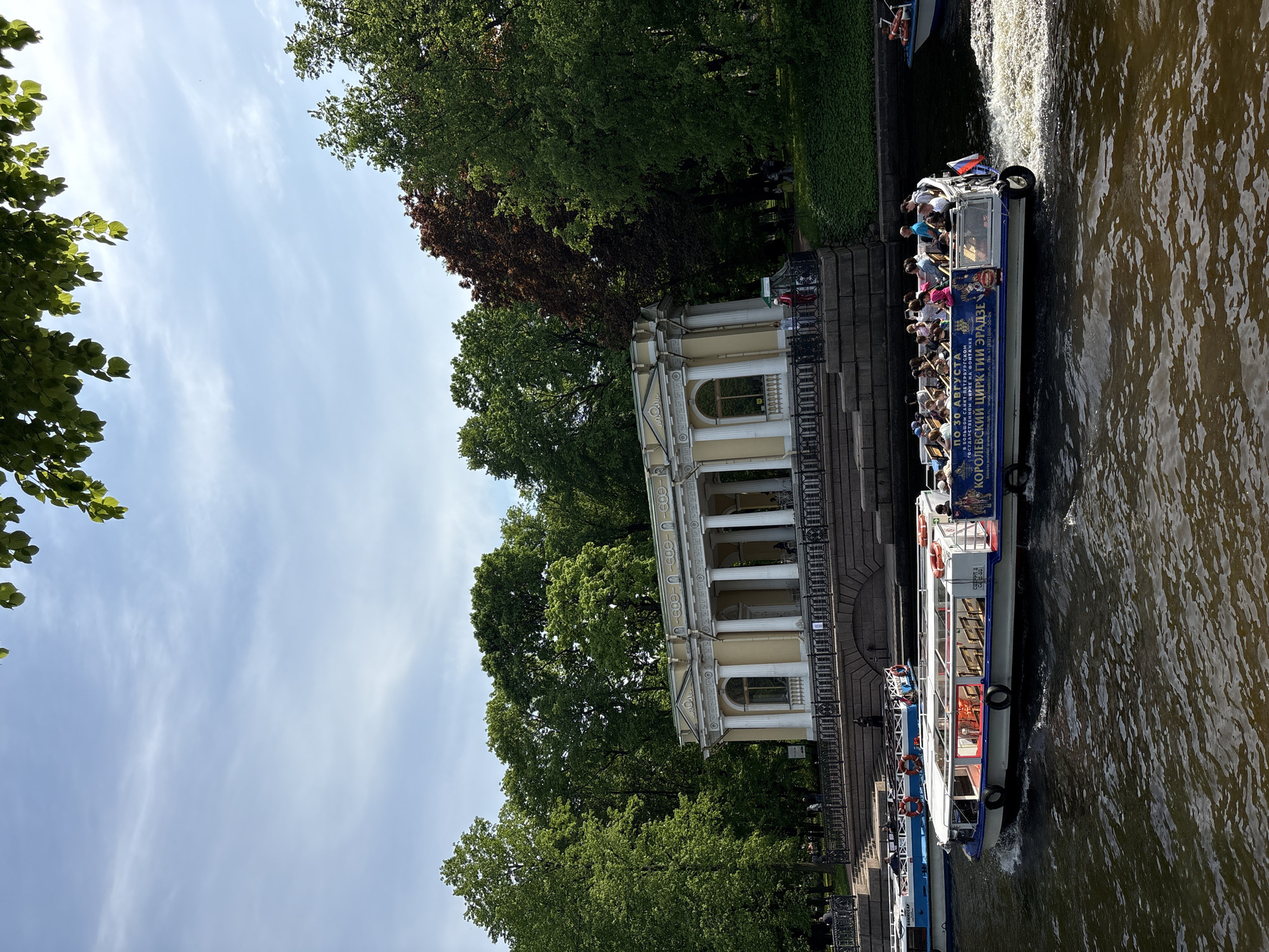 The image depicts a canal scene in Saint Petersburg, Russia, featuring a boat filled with tourists navigating the waterway. The architecture visible includes a classical-style building with columns, positioned next to the canal. Lush green trees surround the building. Sunlight indicates a bright day. The canal's water appears slightly murky. The boat is labeled with "Королевский цирк Гии Эрадзе", indicating a circus tour, and features a Russian flag. Tourists are visible on the boat.